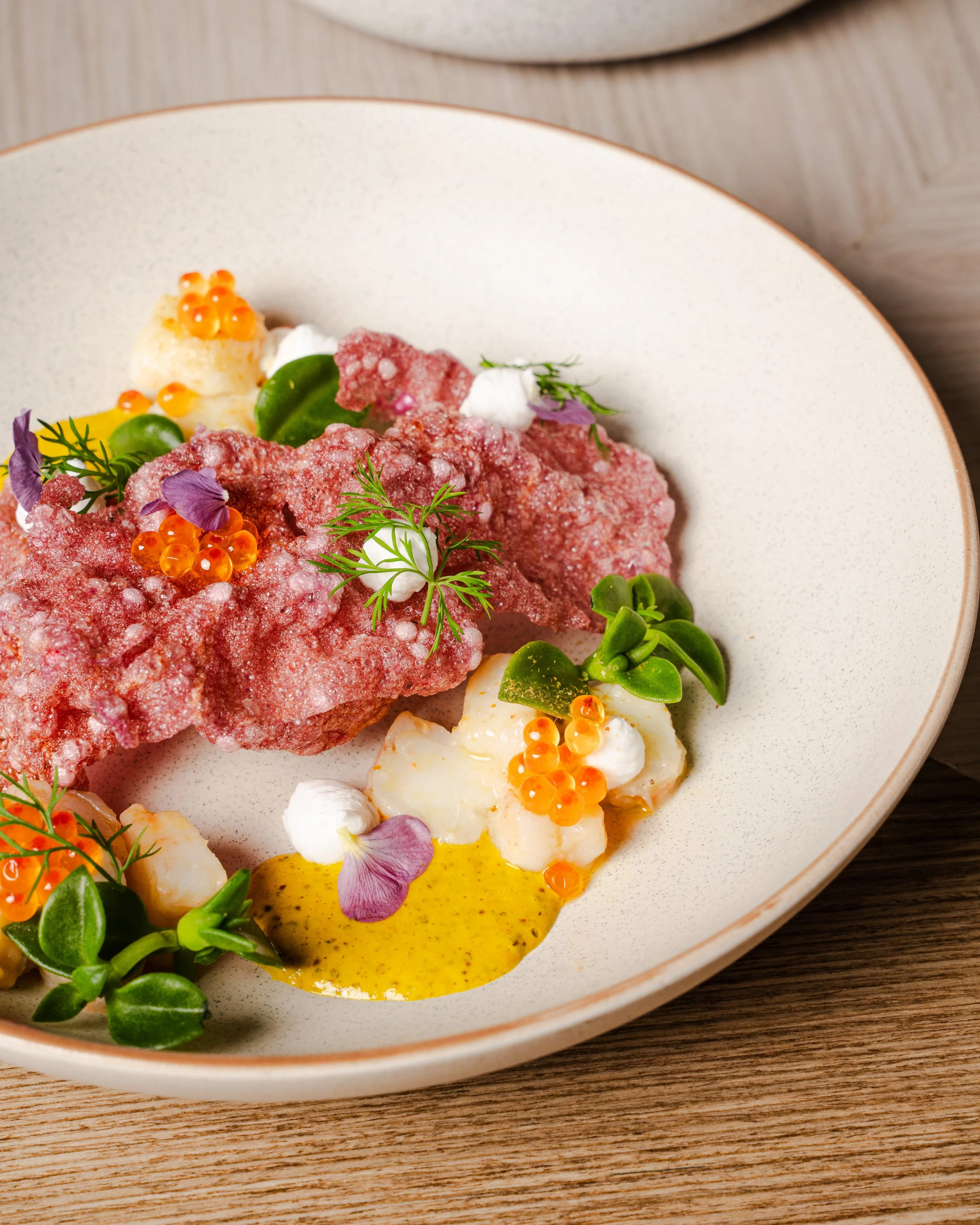 Close-up of a gourmet dish with shredded red meat, green herbs, edible flowers, orange roe, and a yellow sauce on a beige plate with a wooden surface.