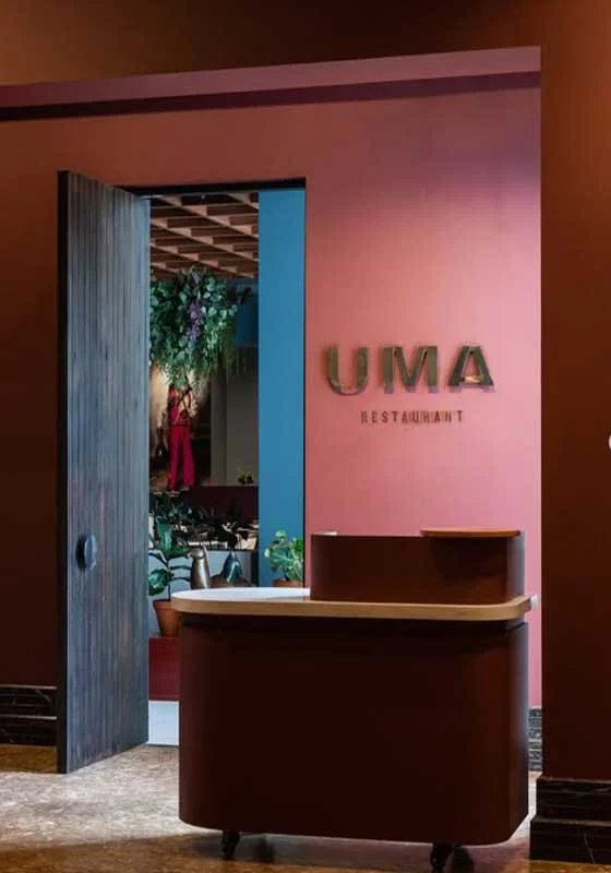 Entrance to UMA restaurant with a wooden door slightly open, pink wall with the restaurant's name, and a reception desk in the foreground.