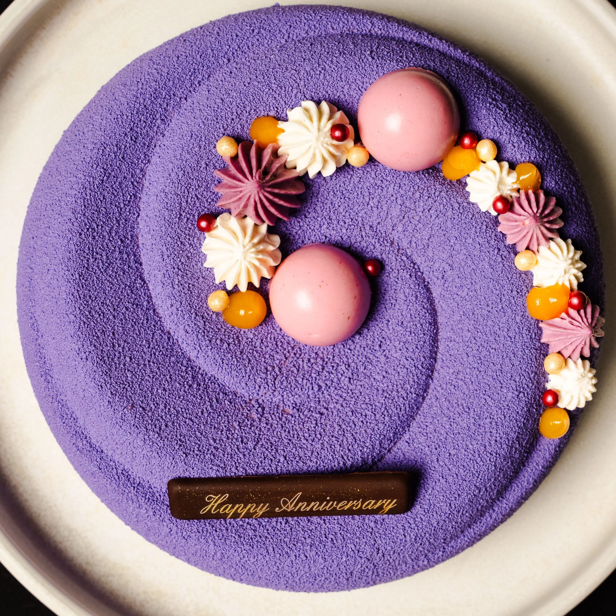 A round purple anniversary cake decorated with pink and white cream dollops, pink and cream spherical decorations, and a chocolate plaque with gold writing that says 'Happy Anniversary'.