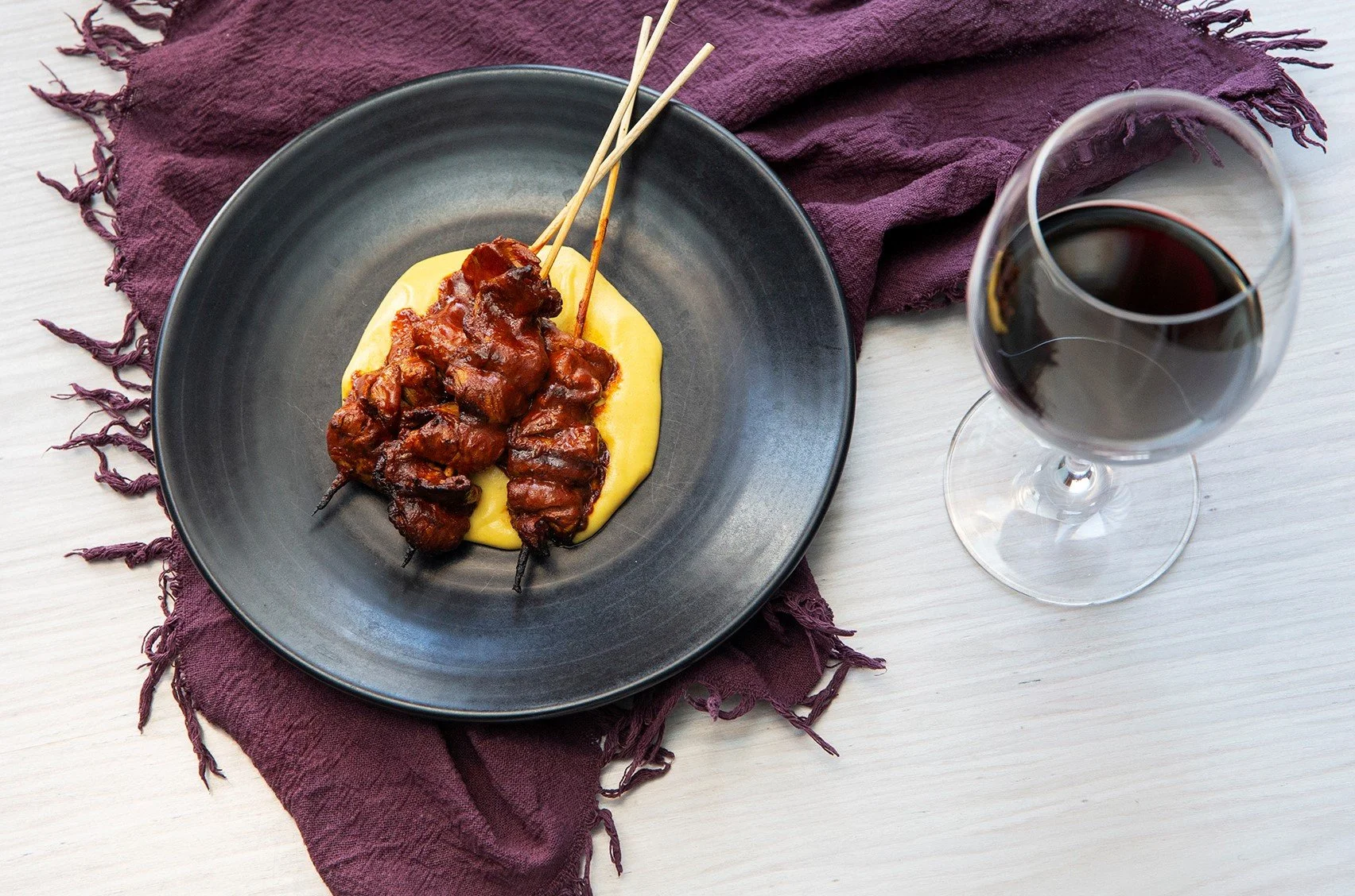 Skewered grilled meat served with mashed potatoes on a black plate, with a glass of red wine nearby and a purple napkin underneath.