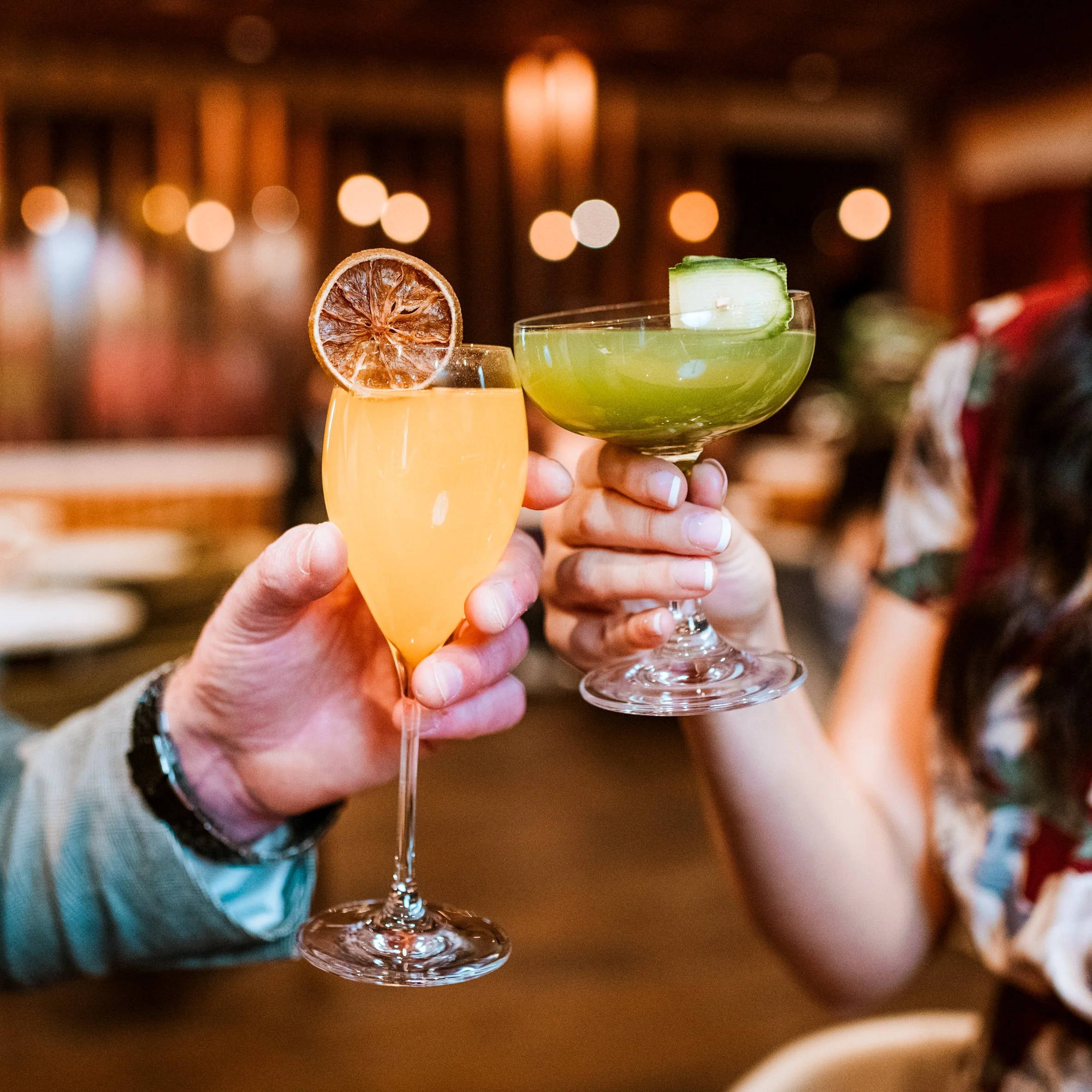 Two cocktail drinks, one orange with a dried slice of orange and the other green with a celery stalk, held in front of a blurred background with warm lighting.