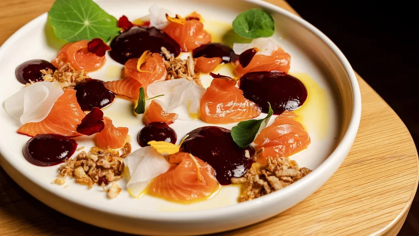 Salmon tartare with dark berry sauce, microgreens, edible flower petals, and crunchy toppings served in a white bowl on a wooden platter.