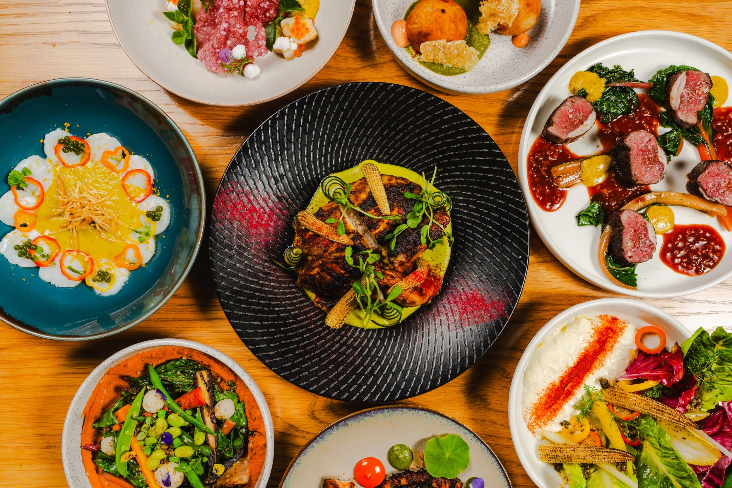 A table set with various dishes including a leafy vegetable salad, a bowl of stewed vegetables, a plate of glazed meat with vegetables, a small dessert, and a glass of red wine.