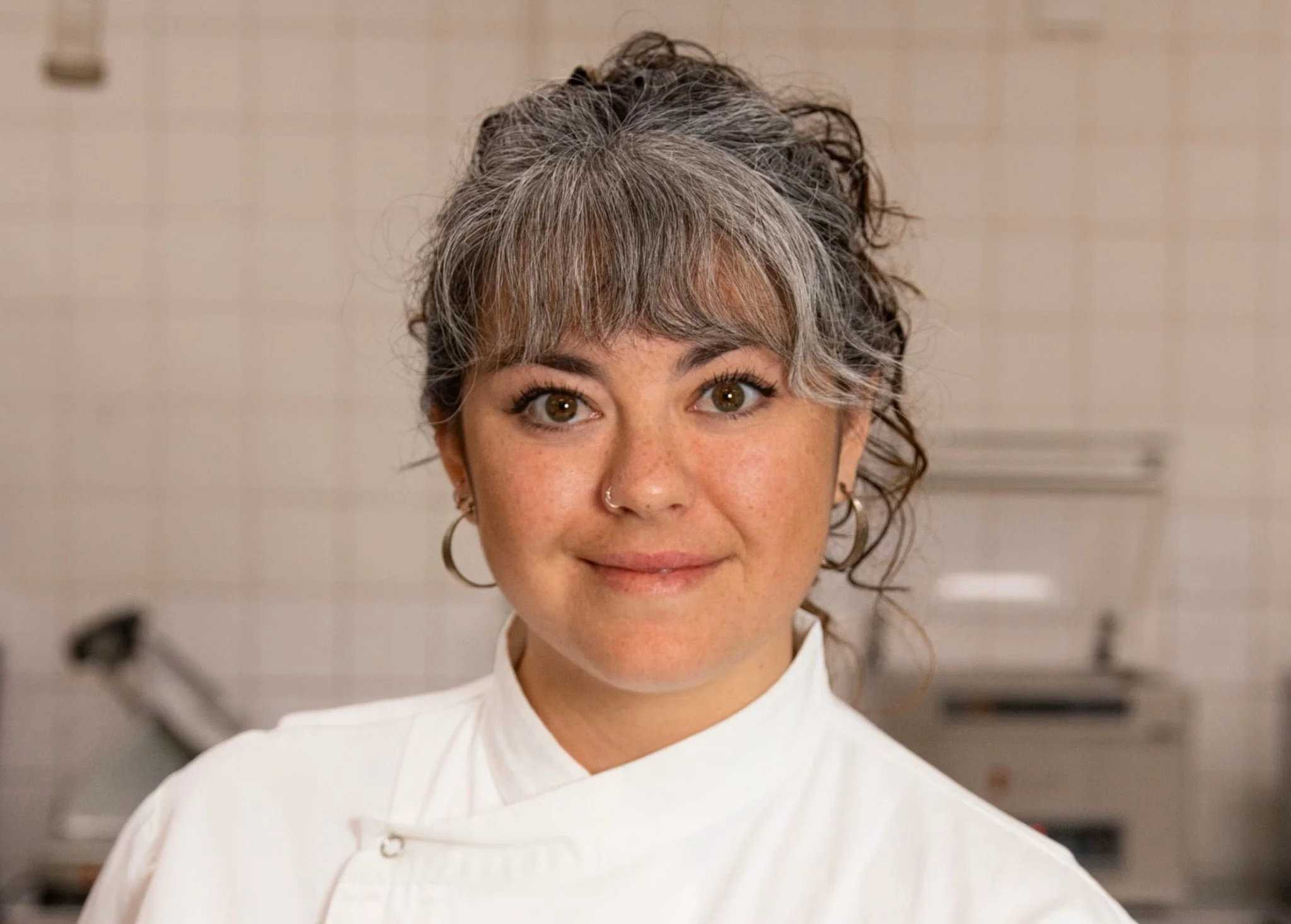 A woman with a mix of gray and dark hair styled in an updo, wearing a white chef's coat, in a professional kitchen setting.
