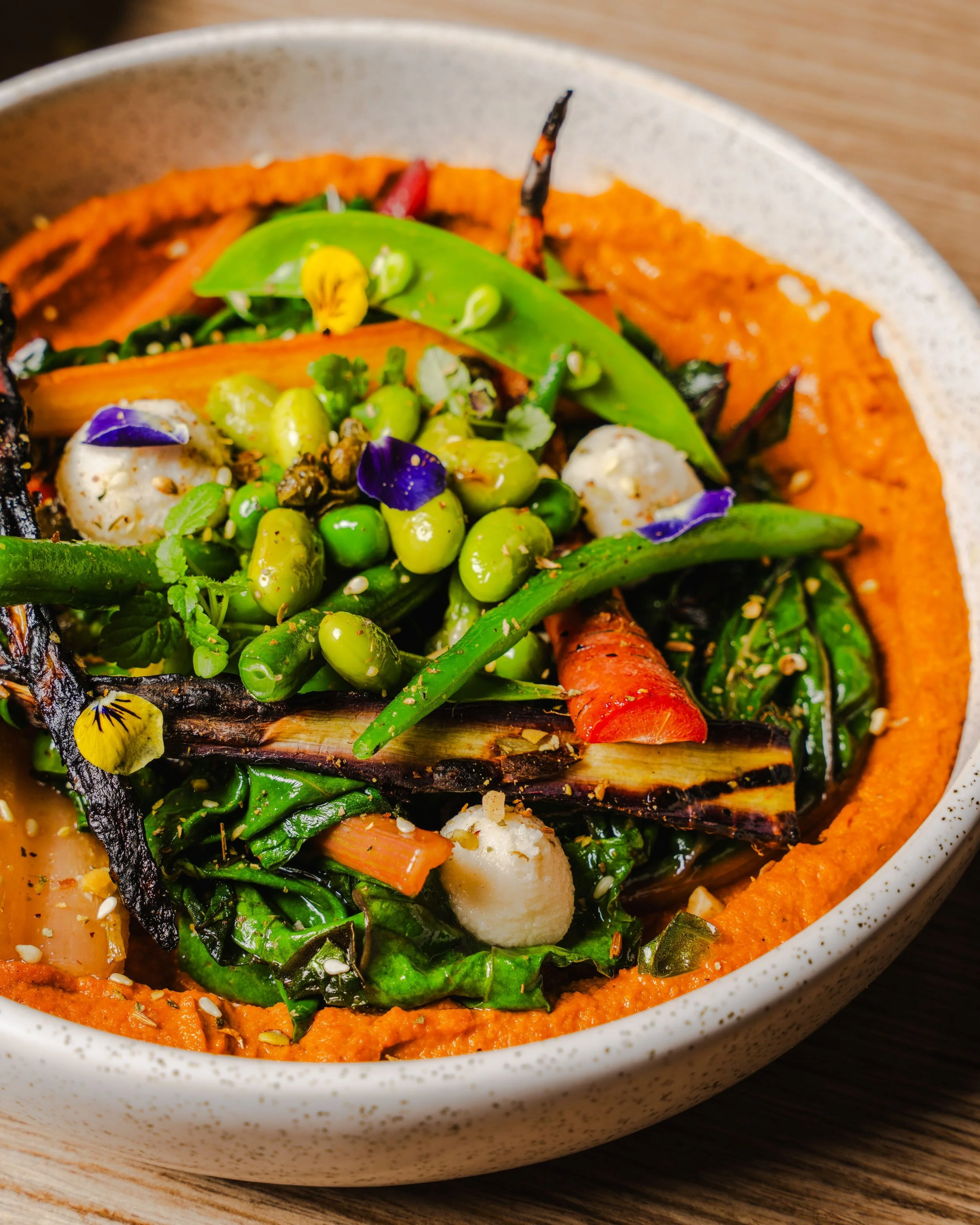 Close-up of a bowl with roasted vegetables and green beans on orange puree, garnished with edible flowers and sesame seeds.