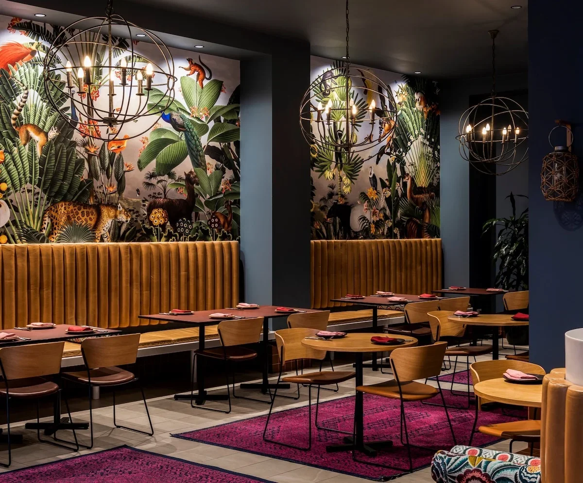 A stylish restaurant interior with jungle-themed wallpaper featuring tropical animals and plants, cheerful wooden chairs, and warm lighting from spherical chandeliers.