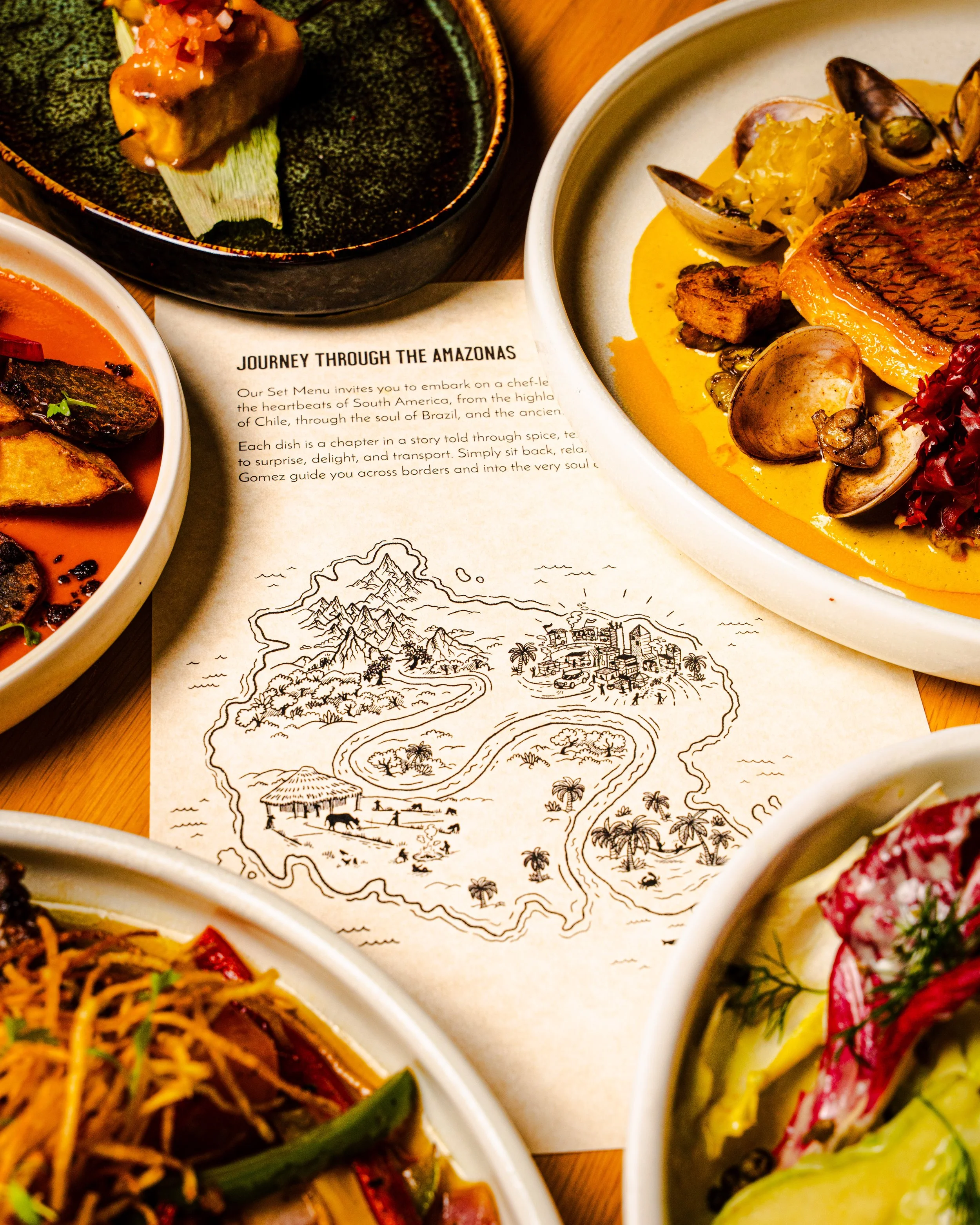 A close-up of an illustrated map showing a route through the Amazon rainforest, with illustrations of mountains, trees, houses, animals, and rivers, surrounded by plates of various colorful dishes of food.