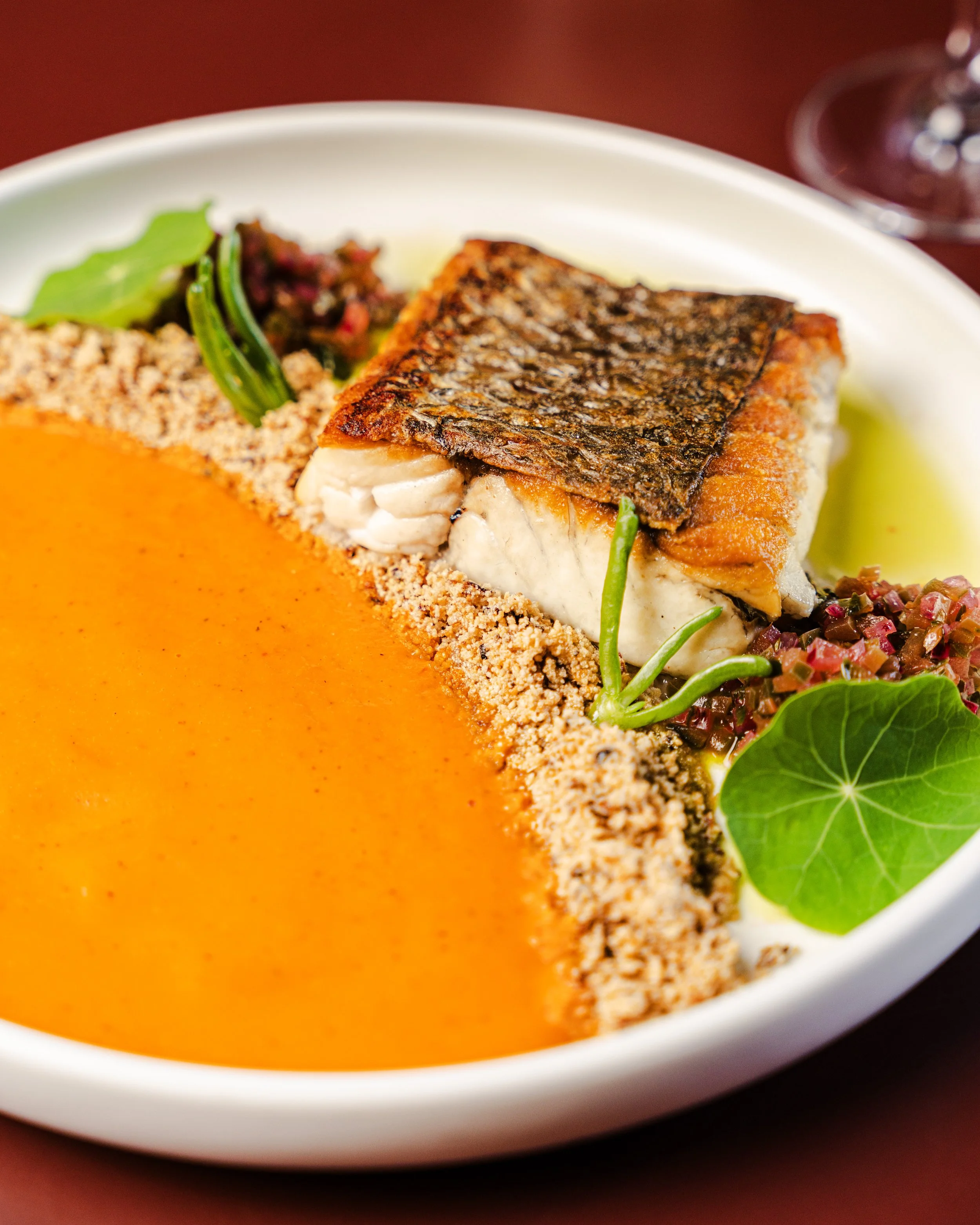 A plate of cooked fish with crispy skin, served with orange sauce, garnished with fresh greens, herbs, and colorful small vegetables.