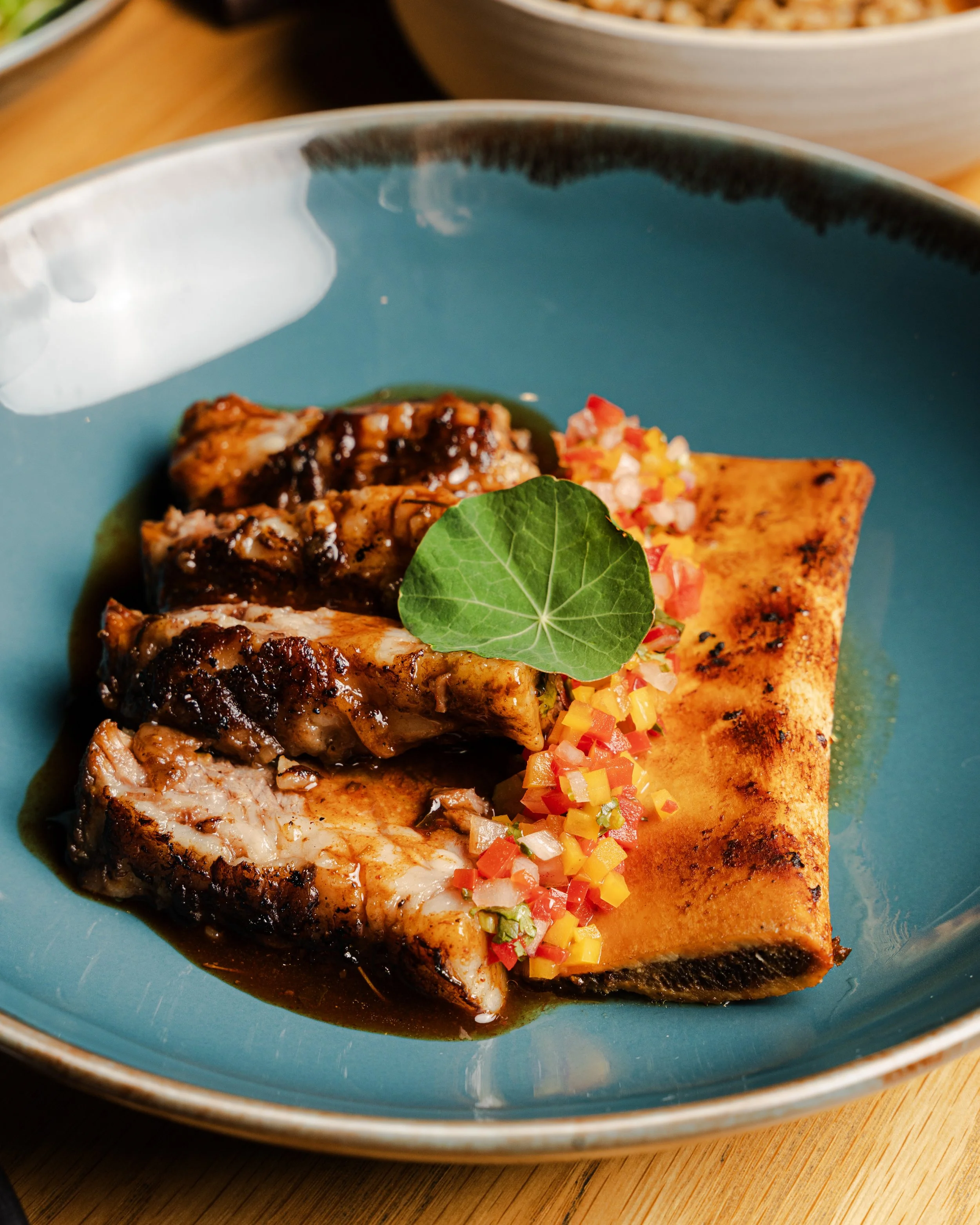 A plate with grilled fish, topped with a green leaf, served with a side of diced colorful vegetables, and a grilled rectangular piece of bread on a blue dish.