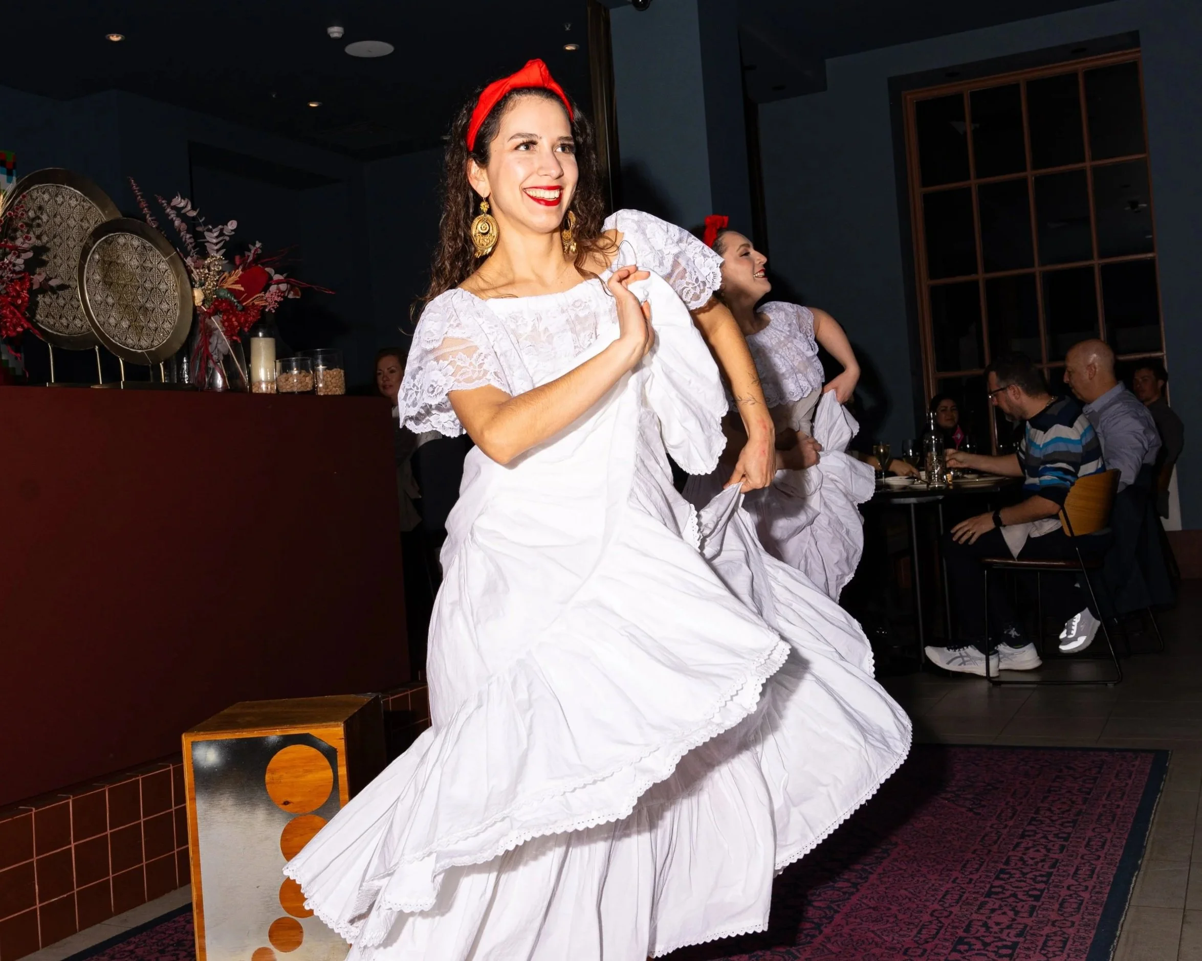 Two women in traditional white dresses with lace details are dancing and smiling at a festive event. The woman in front wears a red headband, gold earrings, and has curly hair. The background features tables with people, floral decorations, and a blu
