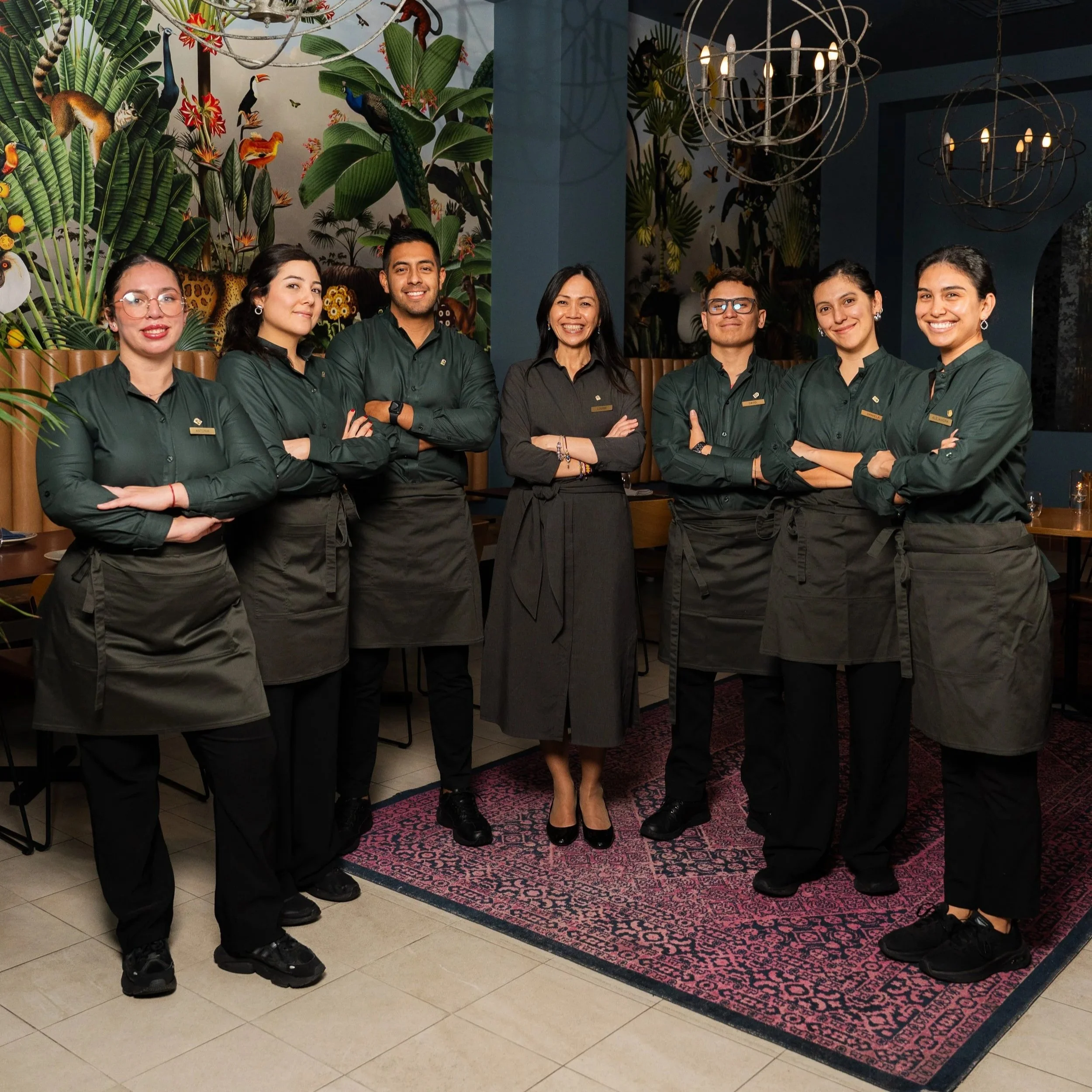 Group of six diverse staff members standing together in a restaurant, dressed in dark green uniforms, smiling and posing for the camera. They are positioned in front of a vibrant tropical-themed mural with lush green plants and colorful animals, under modern chandelier lighting, on a patterned pink and black rug.