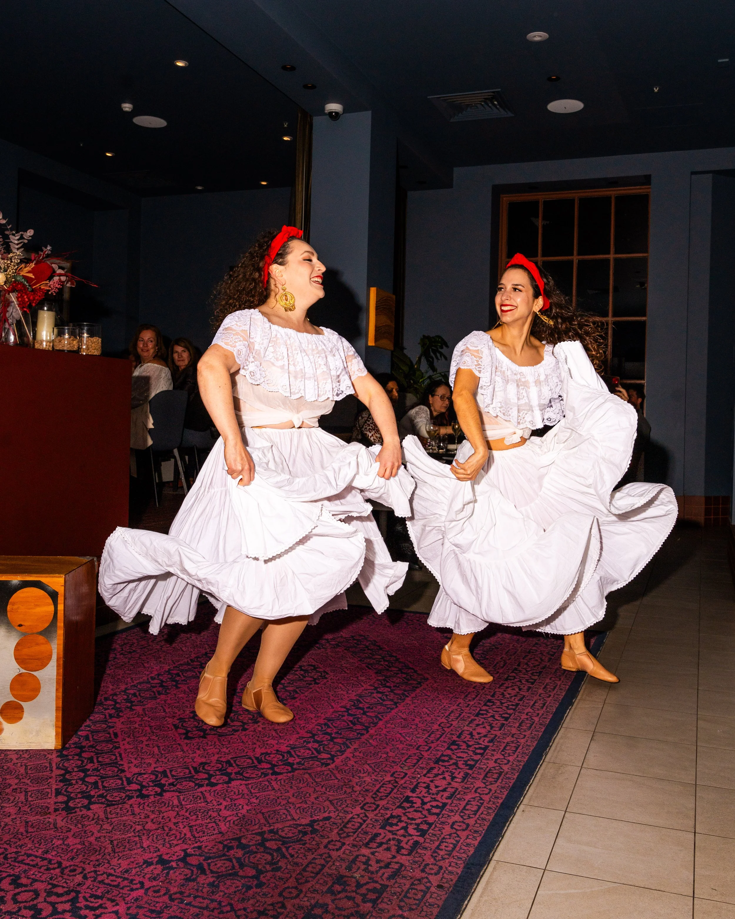 Two women dressed in traditional Spanish flamenco costumes are dancing on a stage, with matching white dresses with ruffled skirts, red headbands, and tan shoes, smiling and enjoying the performance.