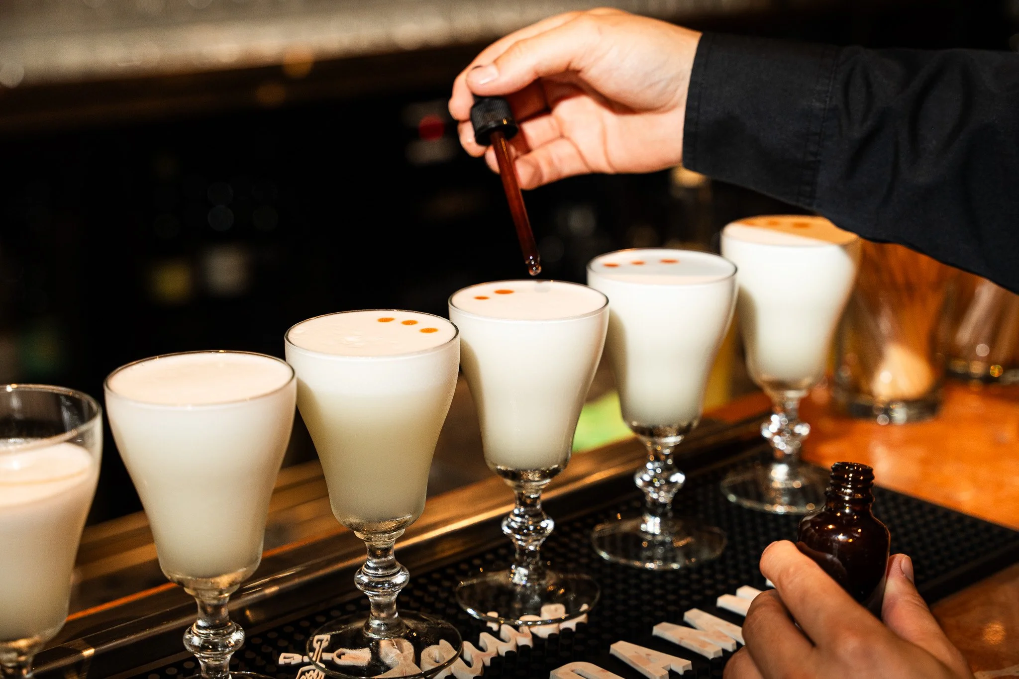 Barista adding drops of bitters or syrup to a row of white cocktail glasses filled with a creamy drink.