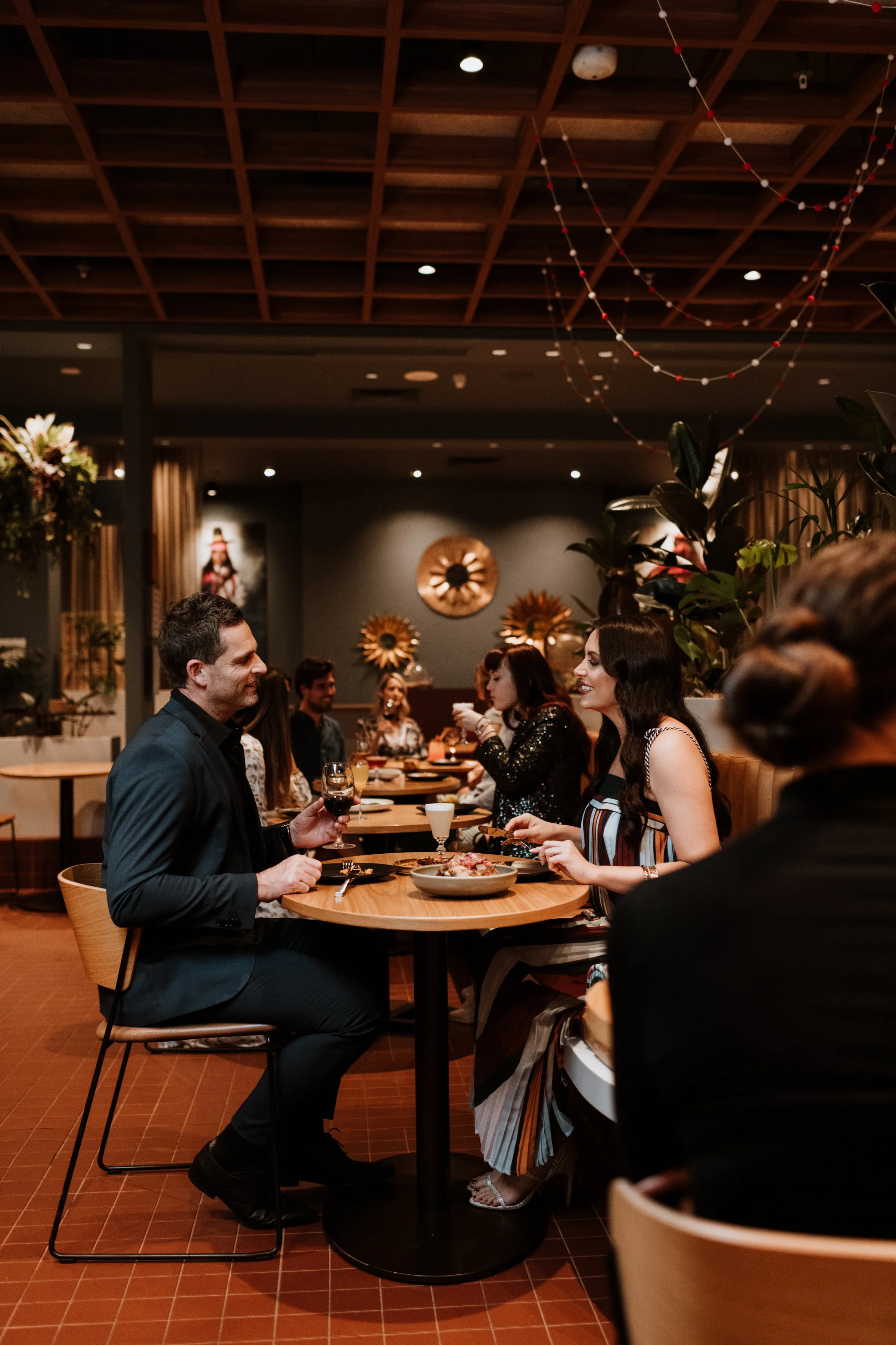 People dining at a restaurant, engaged in conversation, with decorations and plants in the background.
