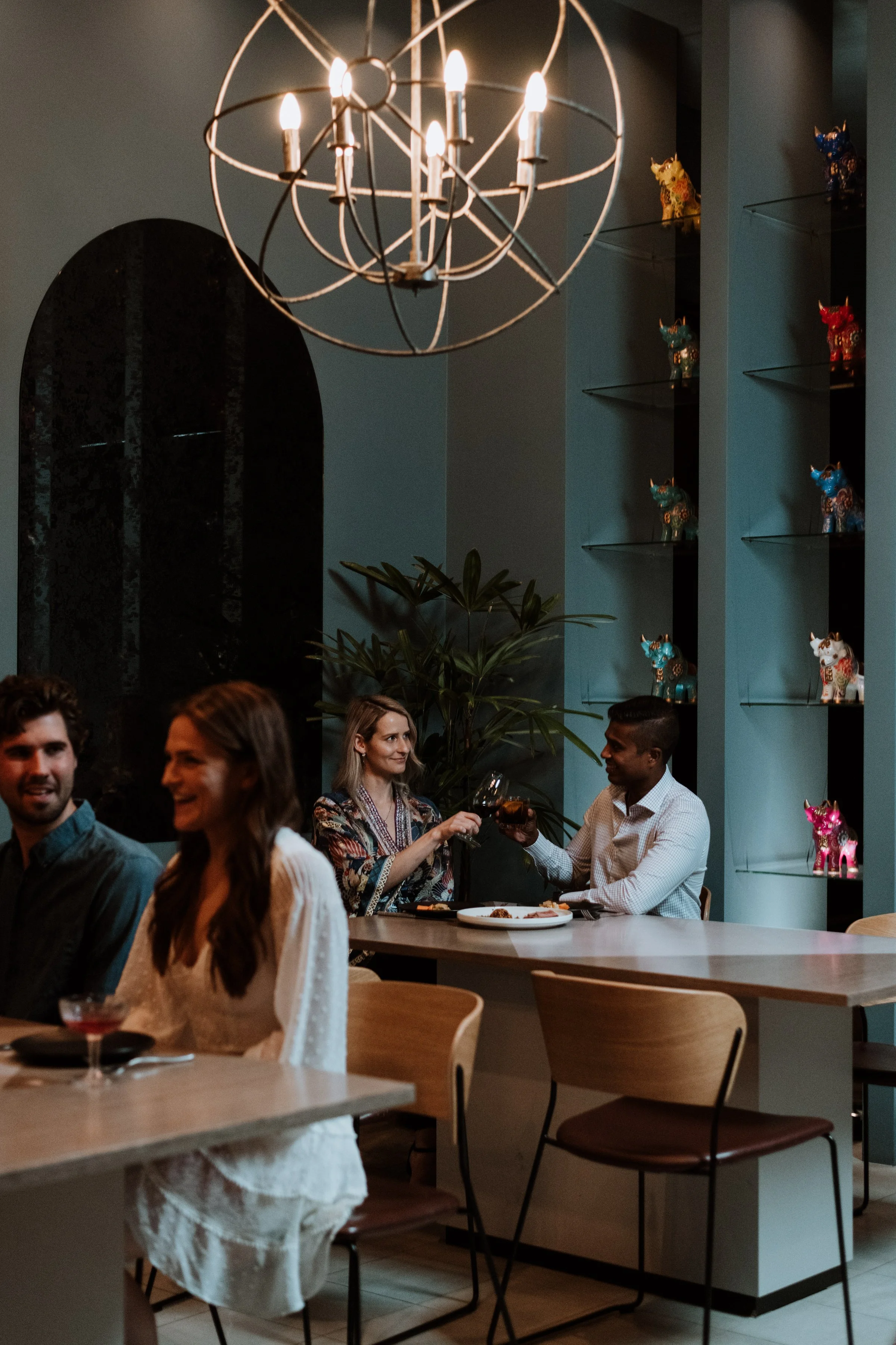 People dining in a restaurant with a modern chandelier and colorful dog-shaped sculptures on built-in shelves.