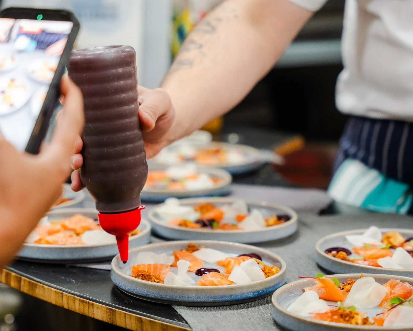 A person is preparing plates of salmon sashimi with garnishes while another person takes a photo of the plates with a phone. Several plates with similar dishes are on a table.