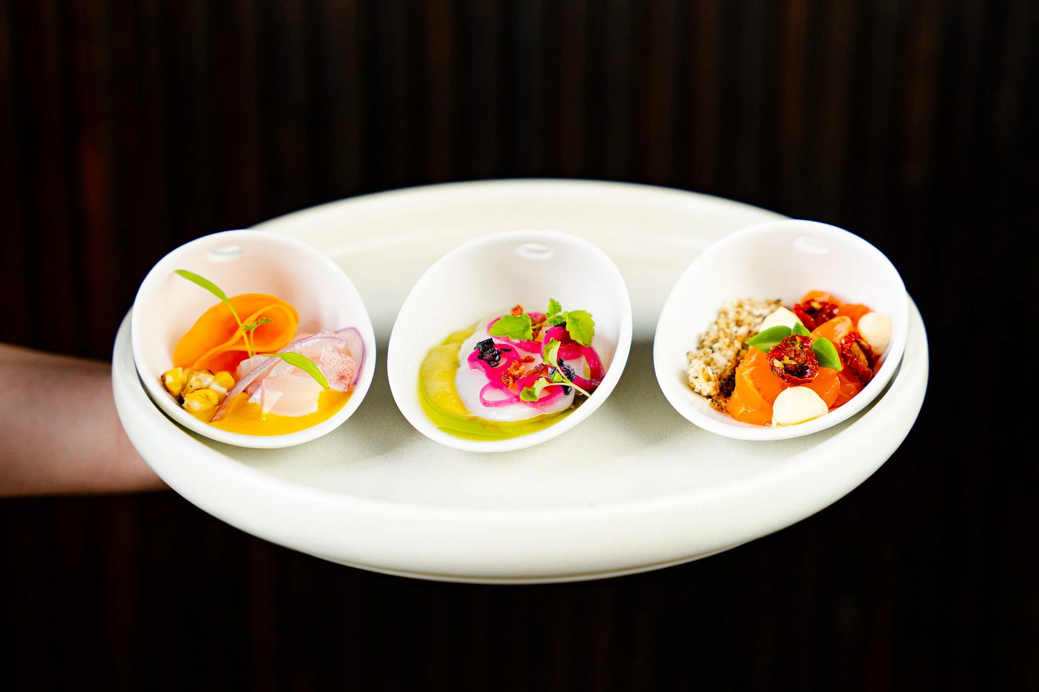 Three small white bowls containing gourmet appetizers on a white tray, with a dark wooden background.