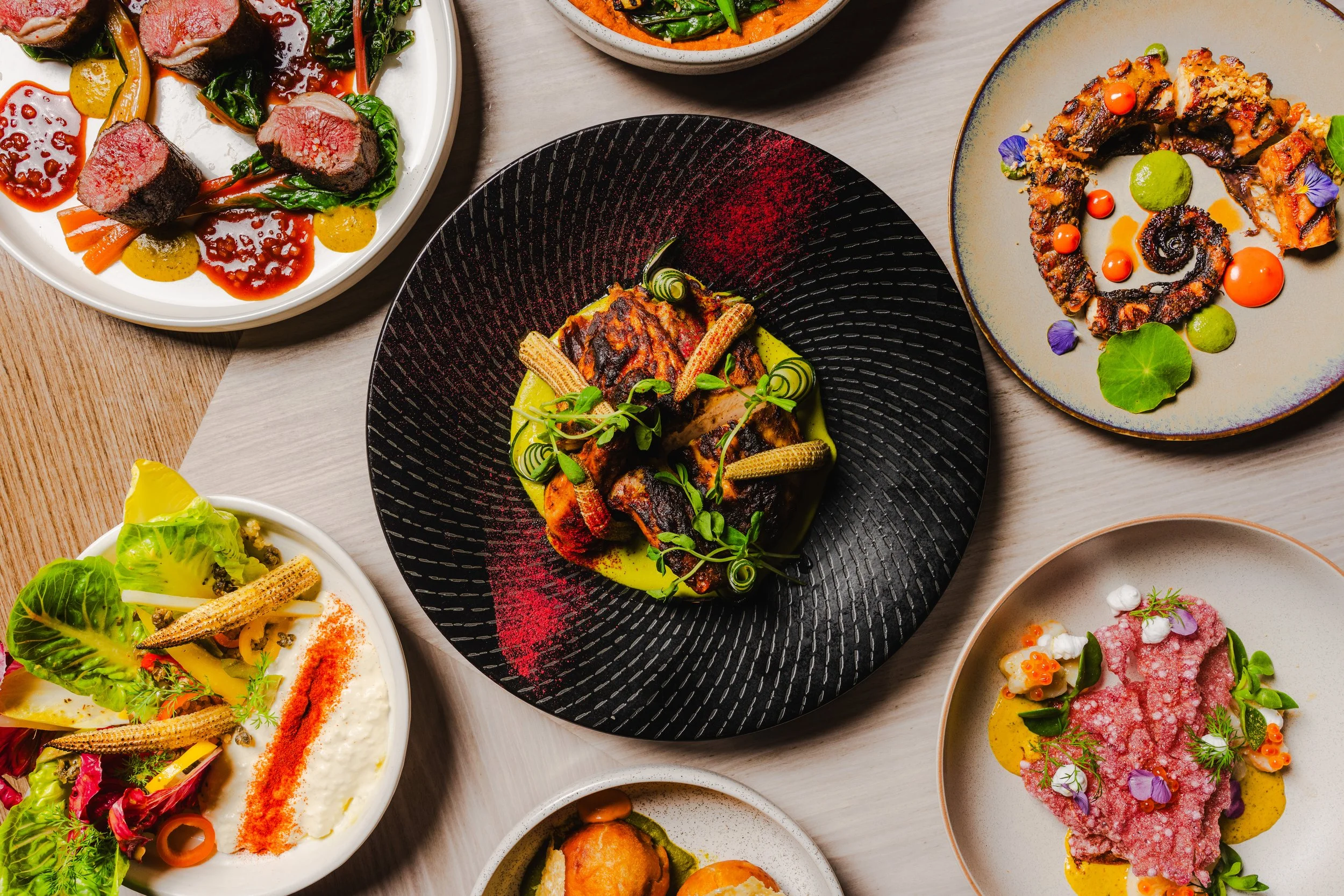 Various plated dishes of gourmet food on a white table, including meat, vegetables, and colorful garnishes.