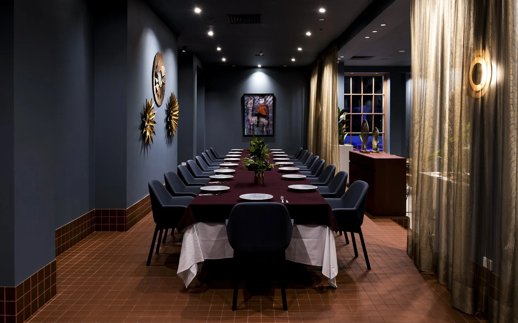 A dining room with a long table set with plates, glasses, and silverware, surrounded by dark chairs, with a dark wall decorated with art and with large windows and curtains on one side.
