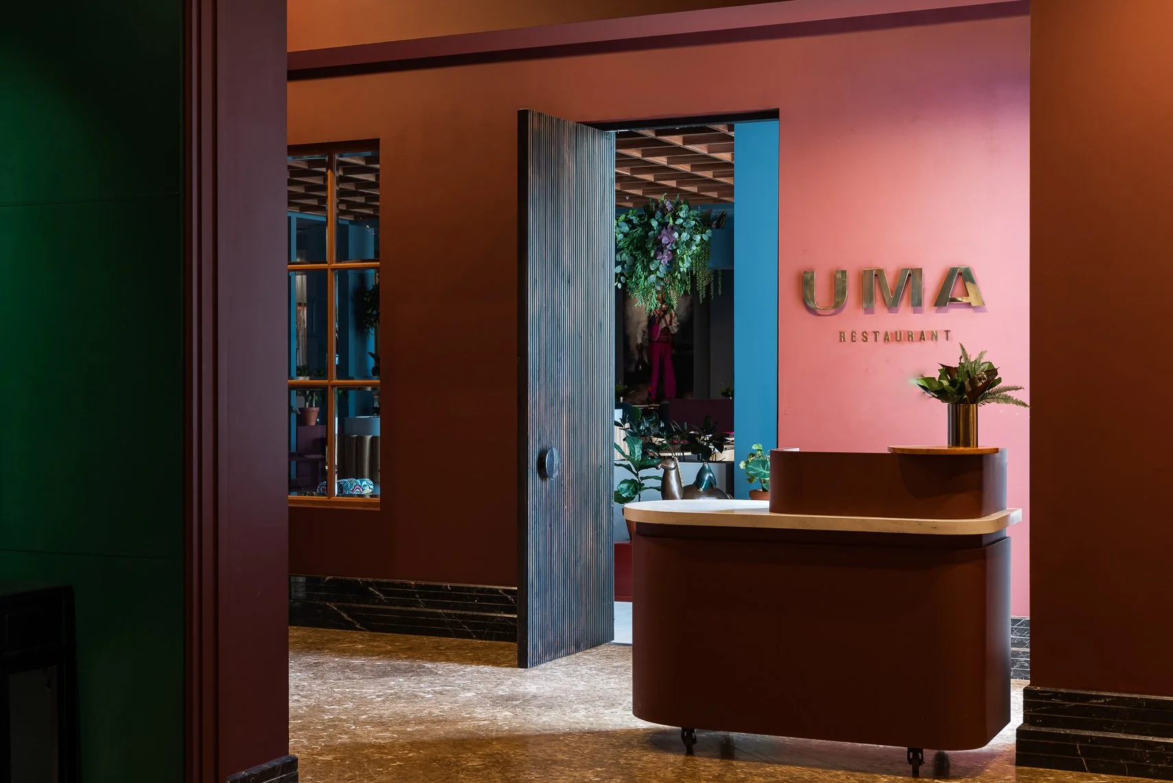 Interior view of UMA restaurant entrance with a pink wall, a gold sign reading 'UMA RESTAURANT,' a curved wooden reception desk with a plant on top, and an open door leading to a decorated dining area with flowers and plants.
