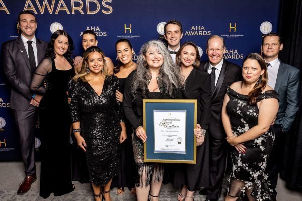 Group of ten people, including women and men, dressed in formal attire, standing on a red carpet in front of a backdrop with awards logos and text, with one woman holding a framed certificate.