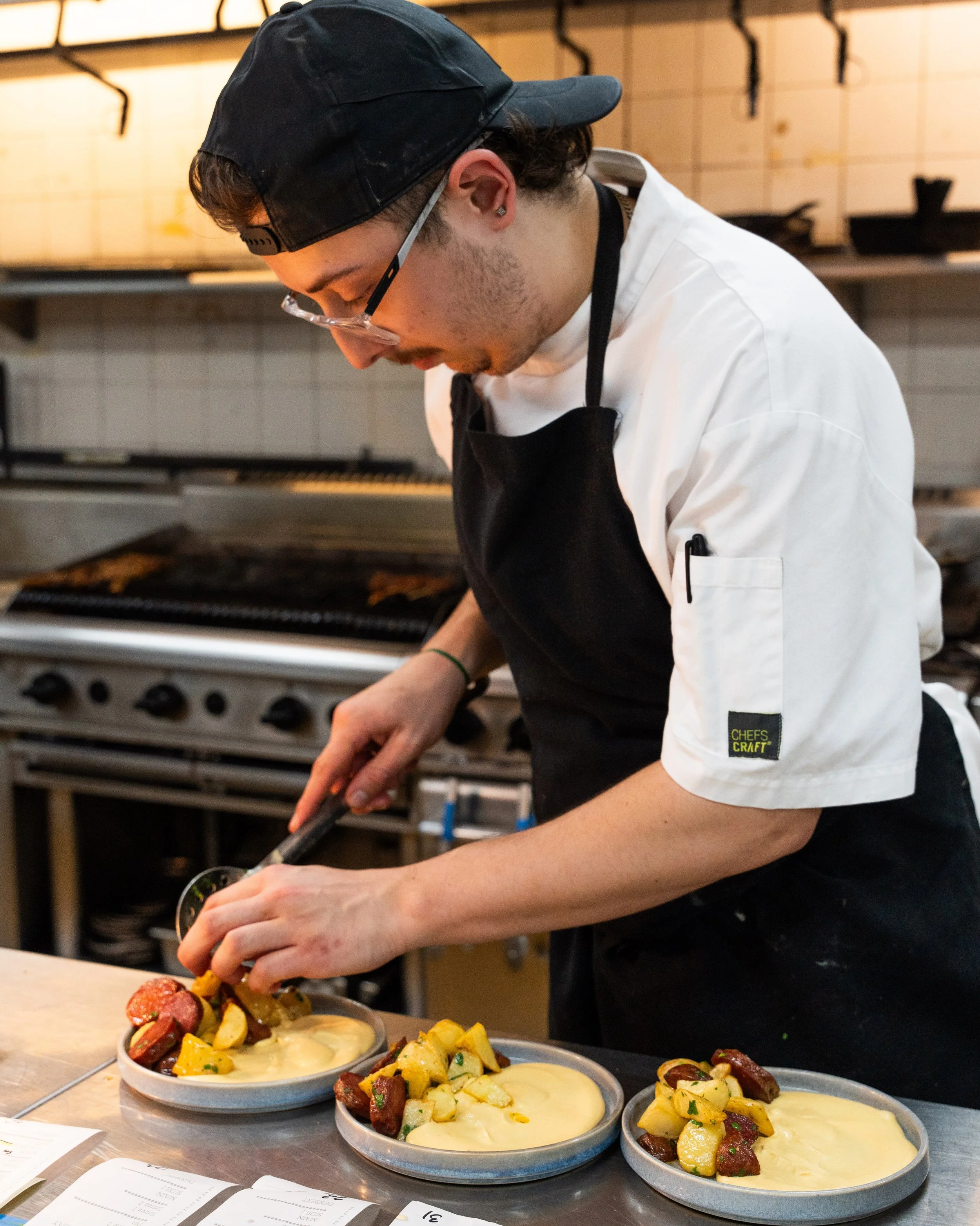 A chef in a white uniform and black apron plate and garnishes multiple dishes of creamy soup with roasted vegetables.