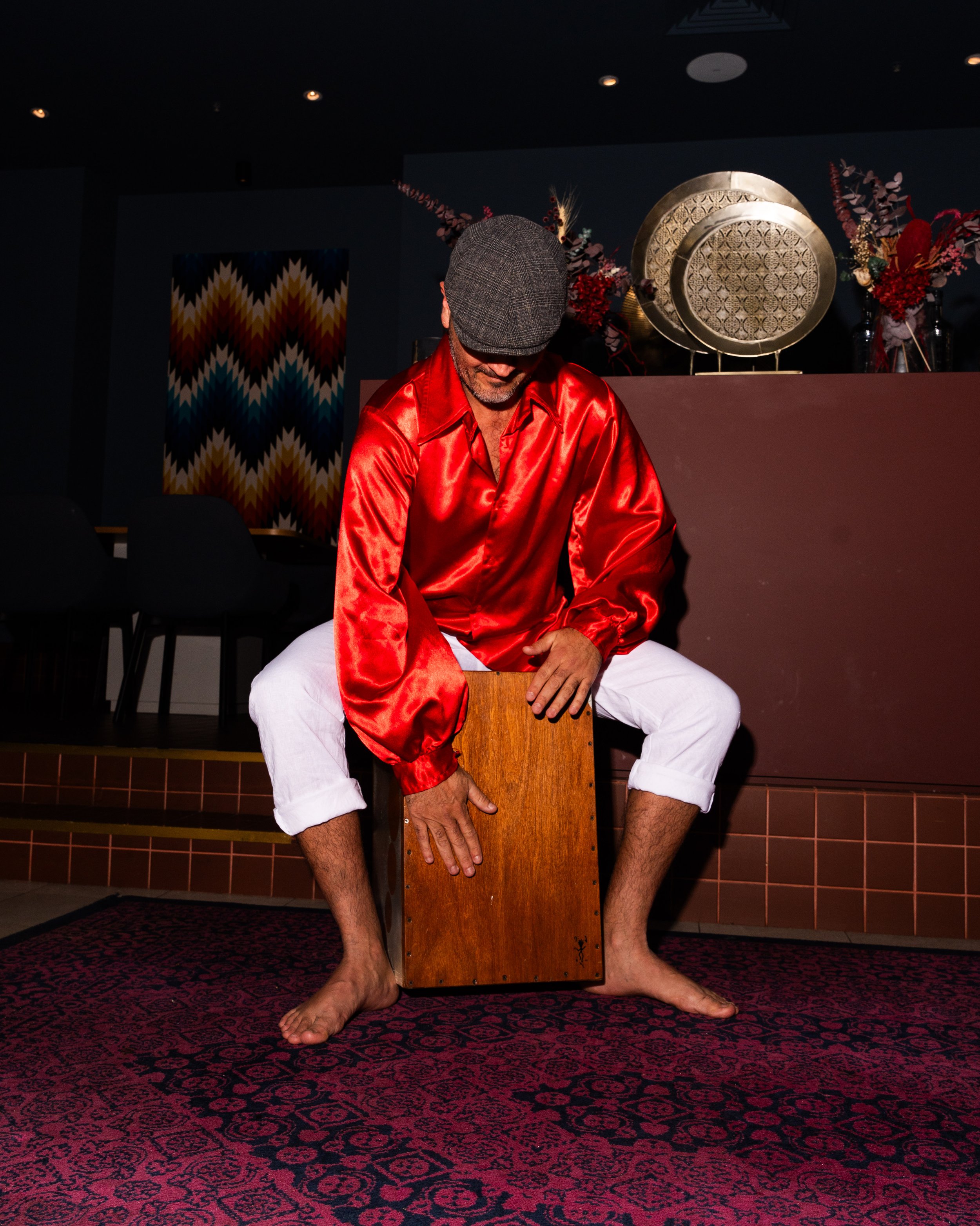 A man with a beard and glasses sitting on a red and black patterned carpet, wearing a gray flat cap, a shiny red shirt, and white pants, holding a wooden box with his hand while perched with one foot on the floor. Behind him are decorative plates, va