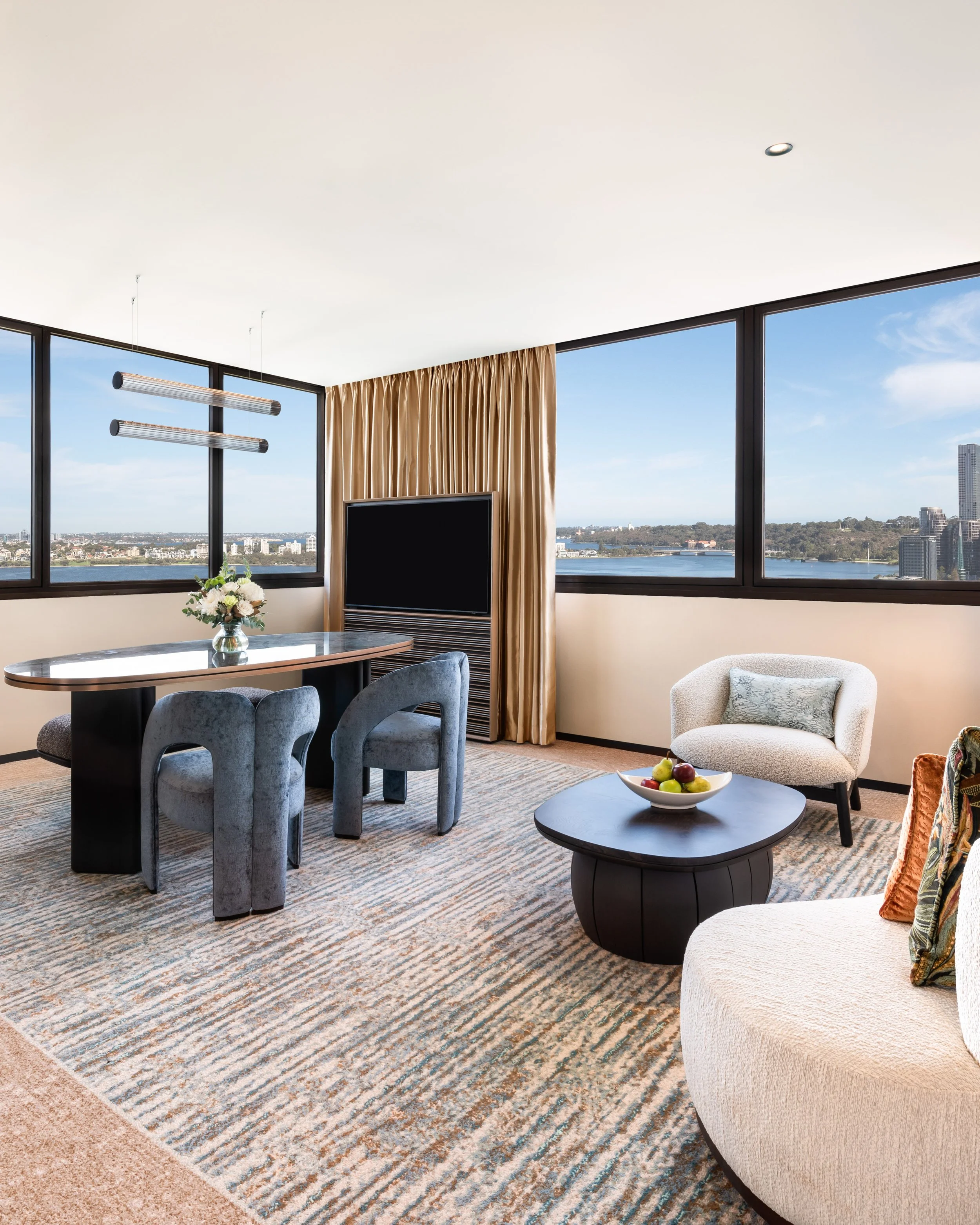 Interior view of a modern living room with large windows showing a cityscape and river, beige curtains, a wall-mounted flat-screen TV, a dark round coffee table with a bowl of apples, a textured cream armchair, and a dining table with floral centerpiece.