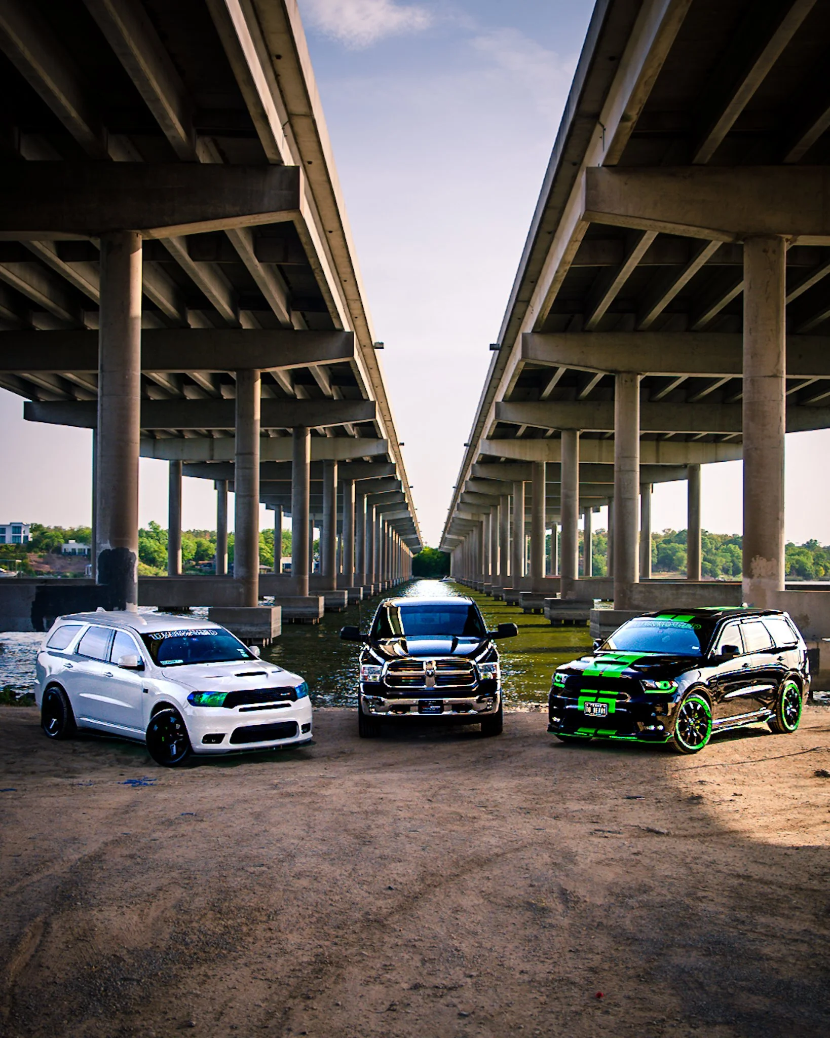 Three cars parked under a bridge with two levels, on a dirt surface, with water and greenery in the background.