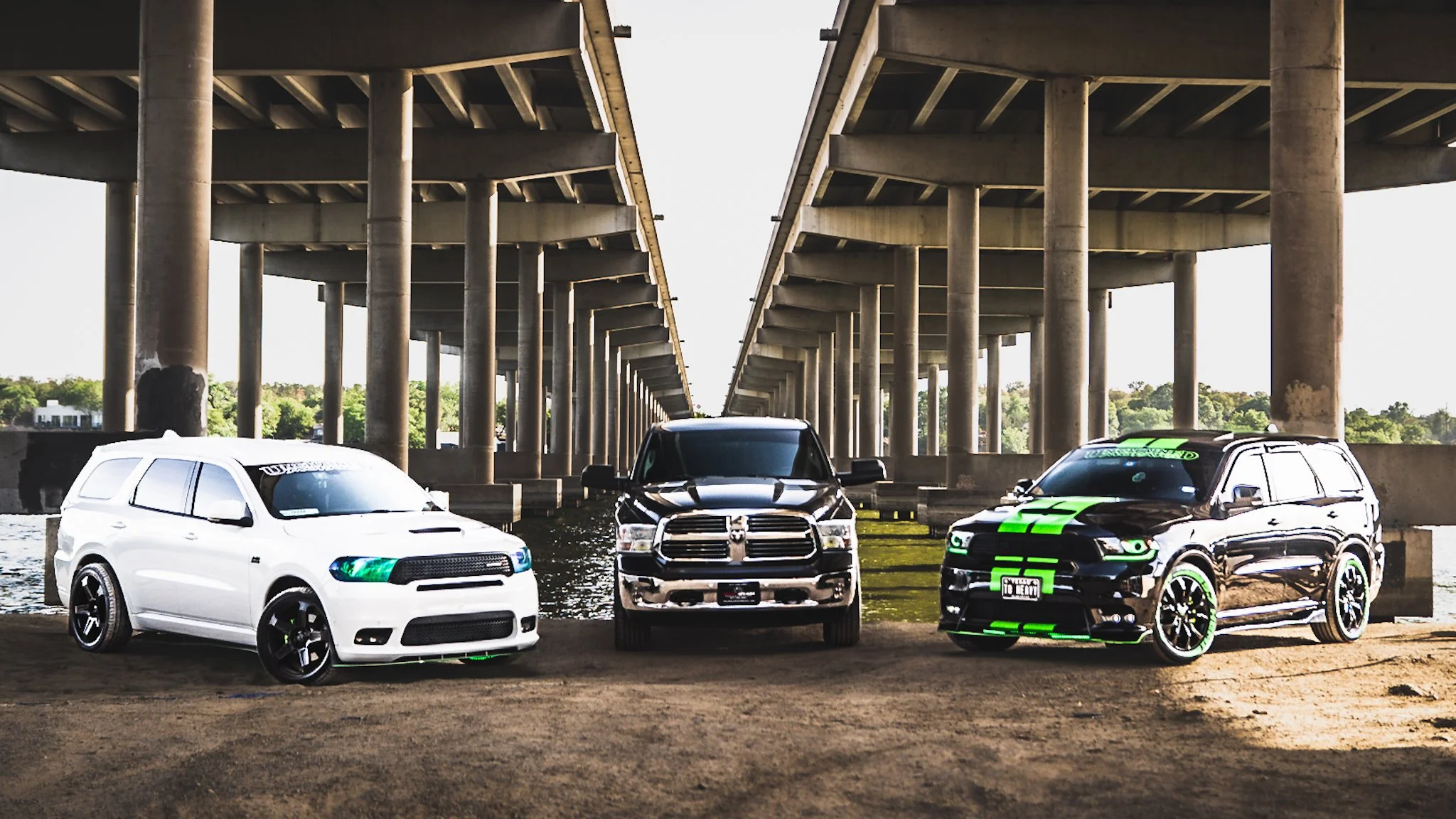 Three pickup trucks parked under a bridge, with water and greenery in the background.