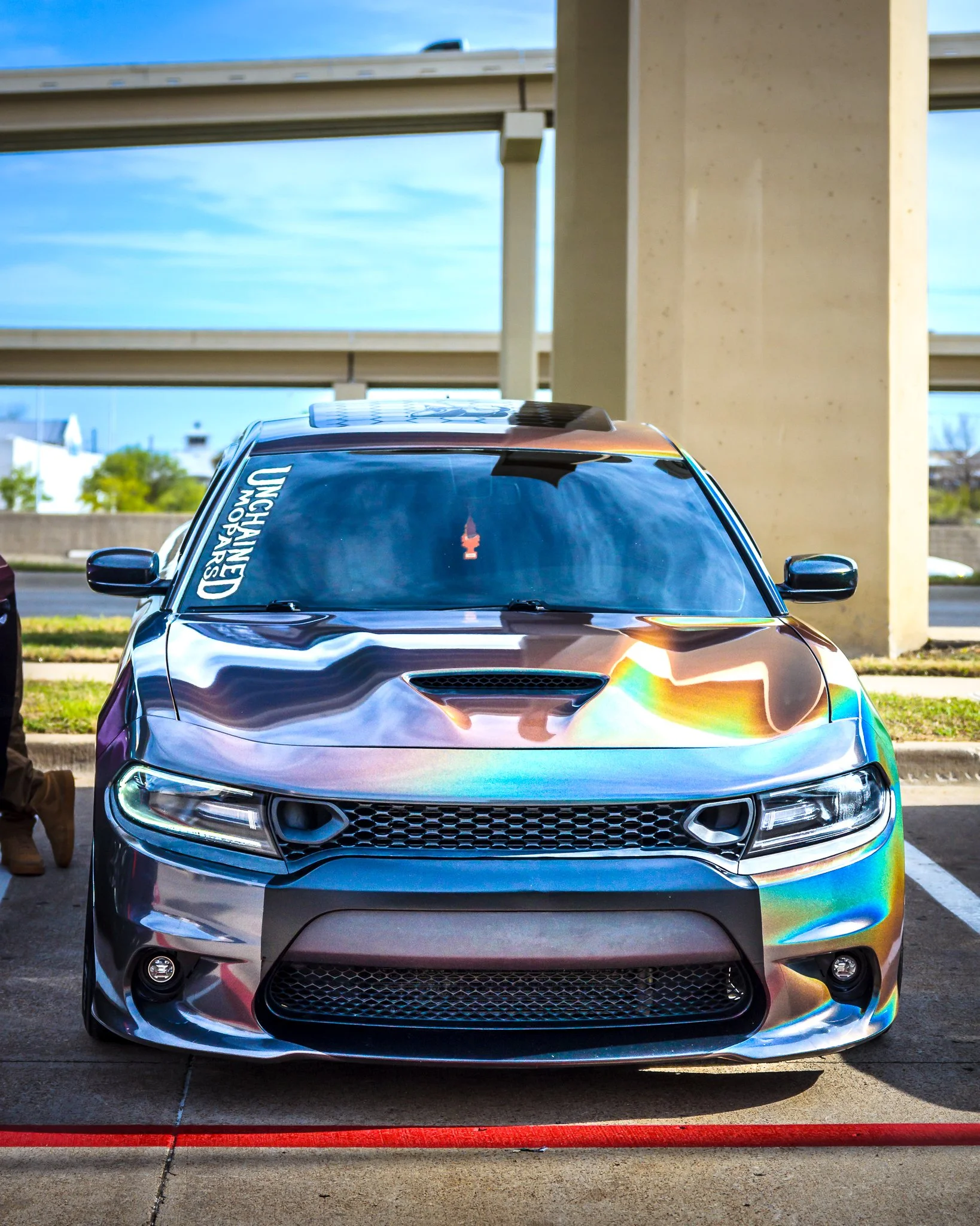 Front view of a shiny, iridescent-colored car with a 'Unhinged Motor's' sticker on the windshield, parked under an overpass on a sunny day.
