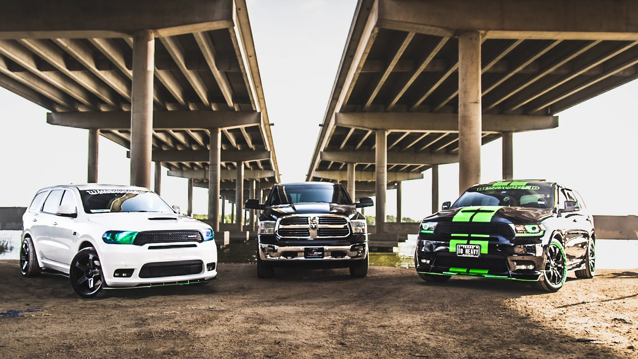 Three modified trucks parked under a bridge, with a white truck on the left, a black truck in the middle, and a black and green truck on the right.