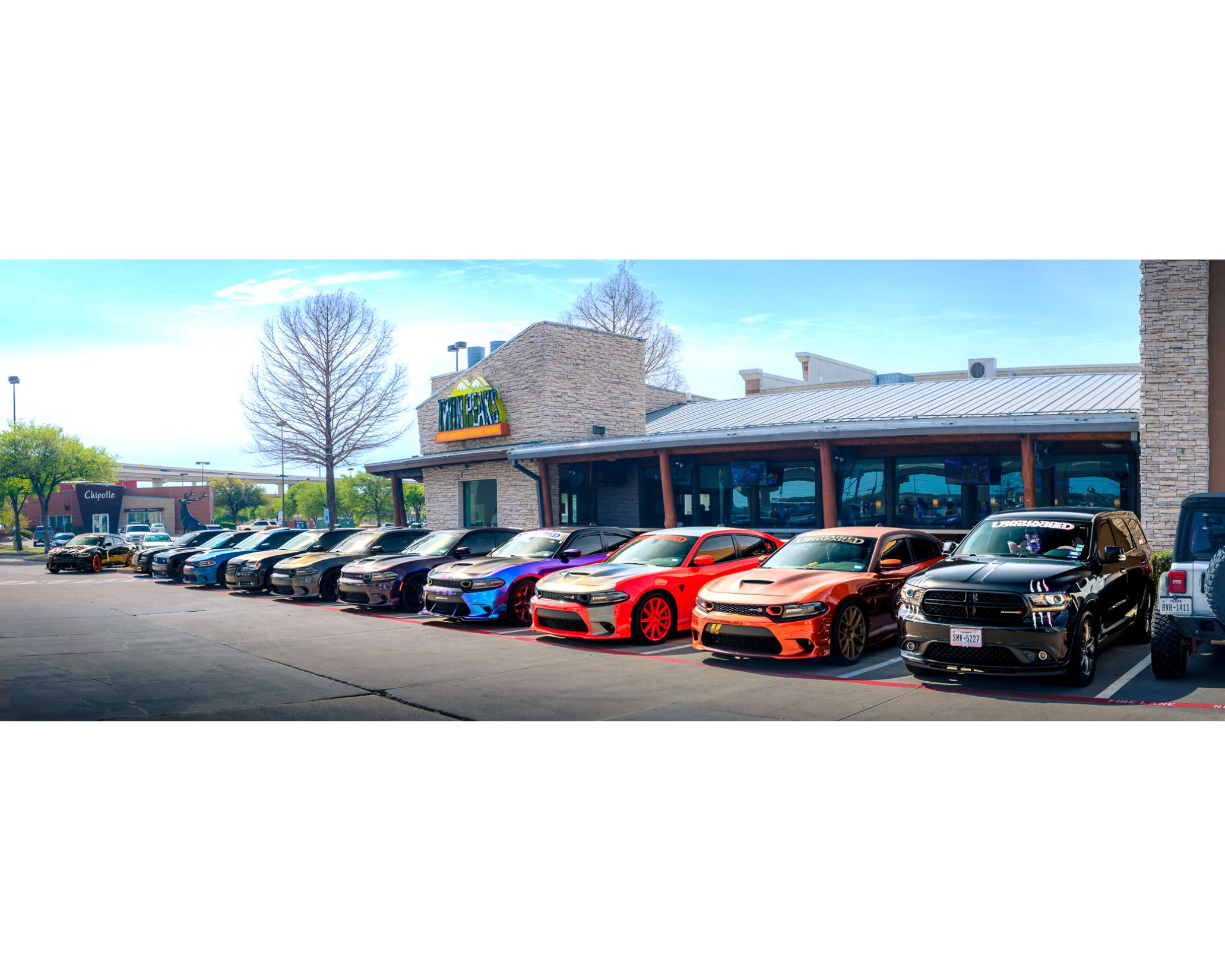 A lineup of various sports cars parked outside a restaurant with a stone facade and a sign that reads 'The Alley' in a shopping area.