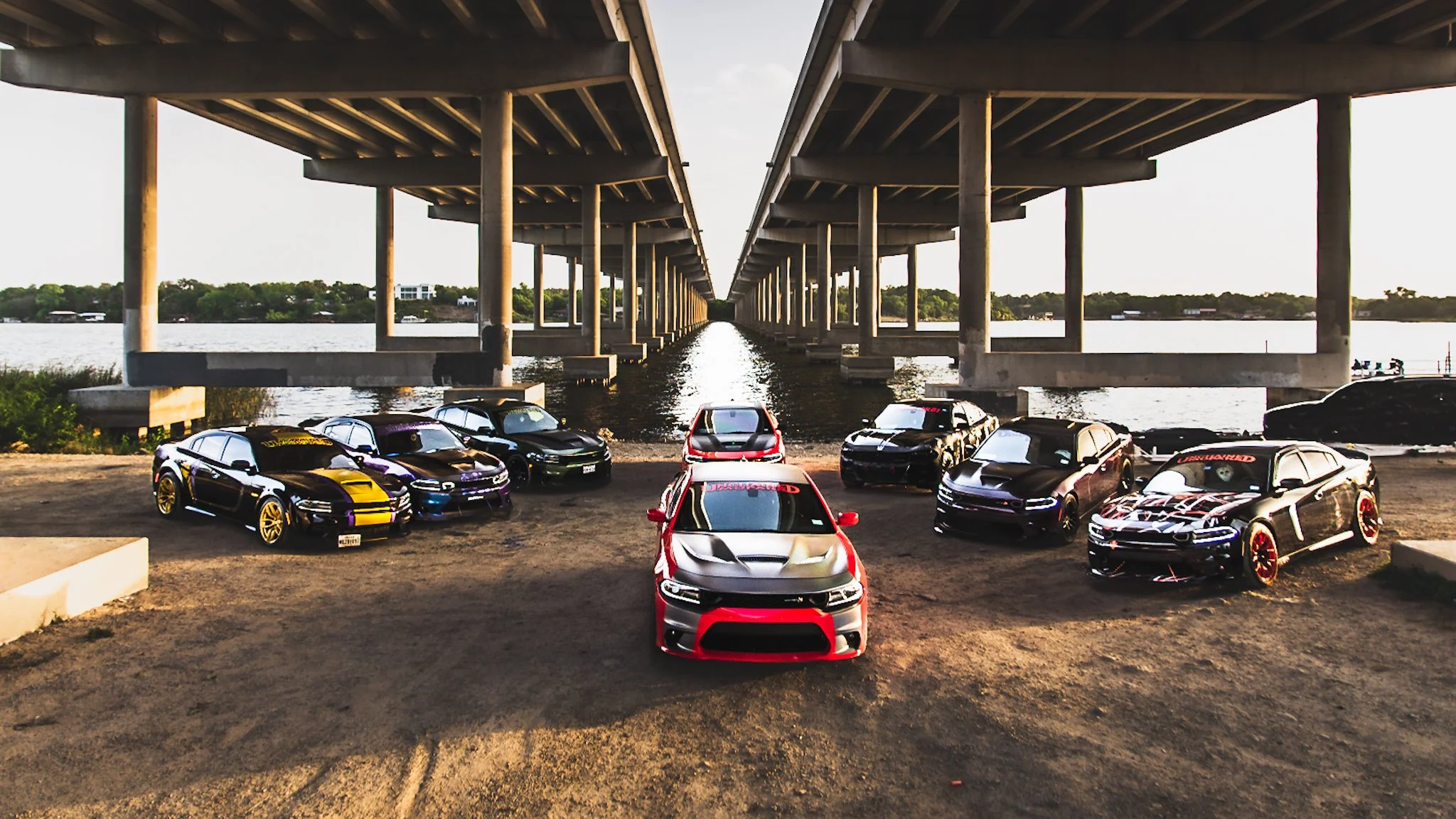 Multiple customized cars parked under a bridge over water at sunset.