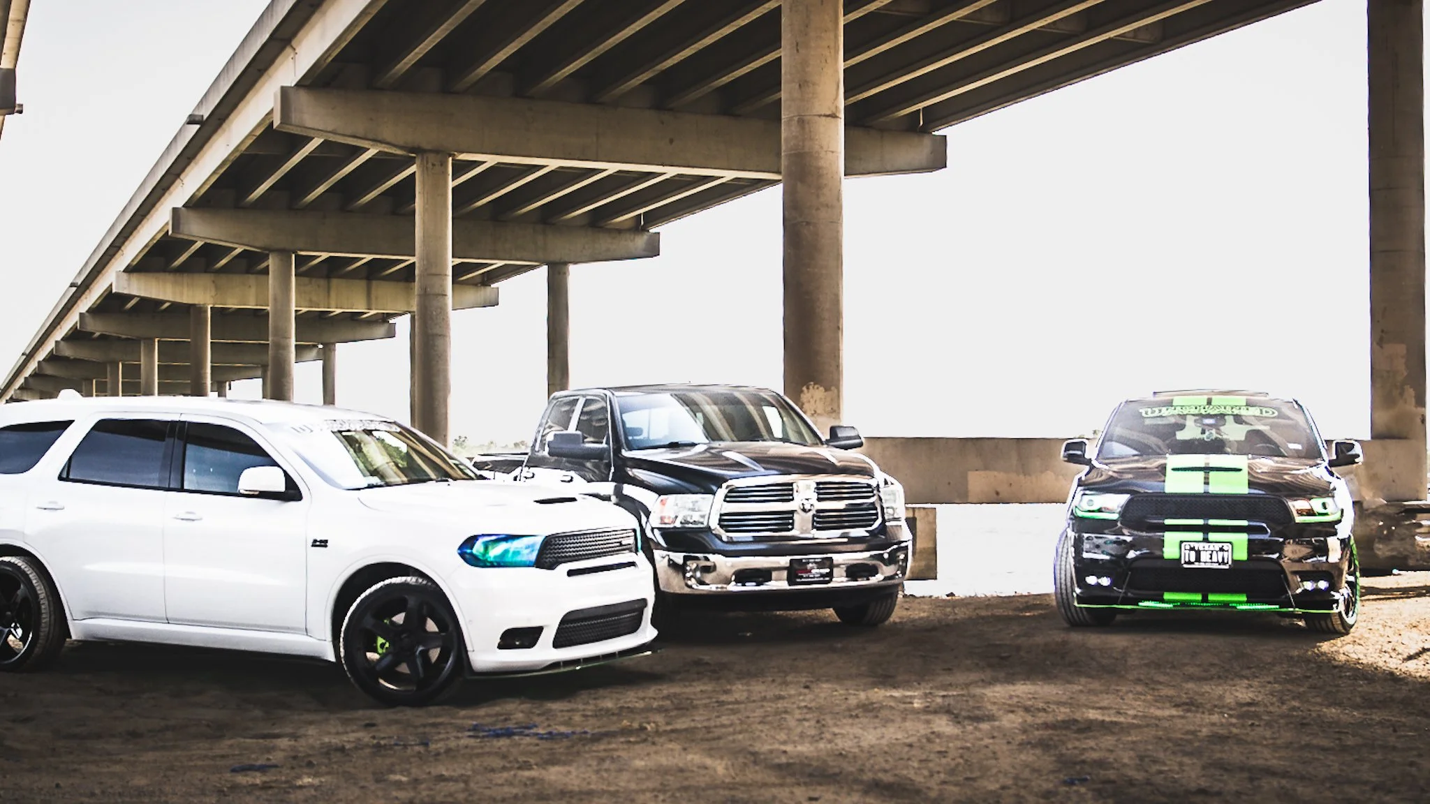 Three cars parked under a bridge, with the middle black pickup truck in front and two sedans on either side, one white and one black with neon green accents.