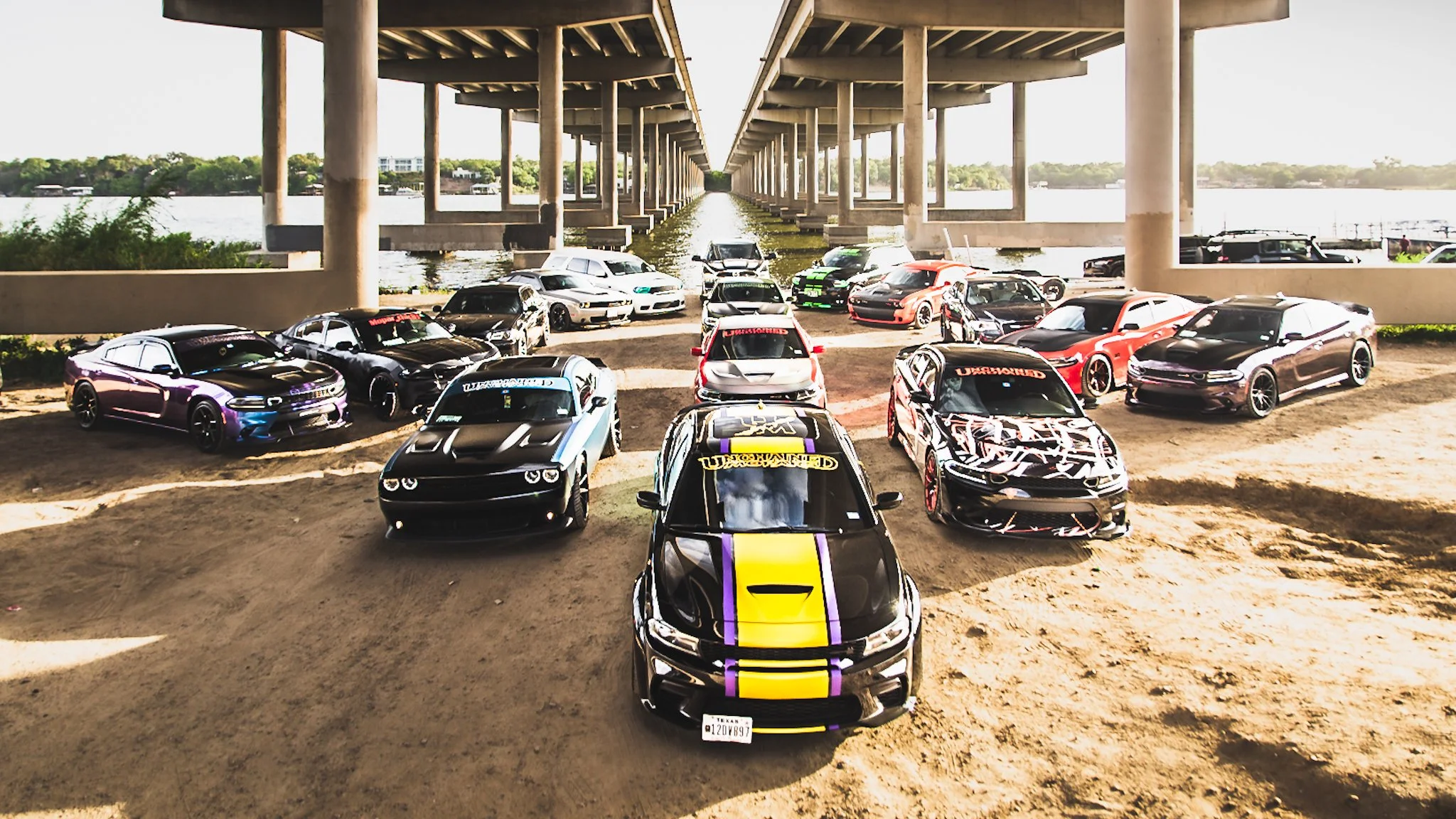 A group of modified sports cars parked under a bridge near a body of water, with the cars arranged in a semi-circle facing forward.