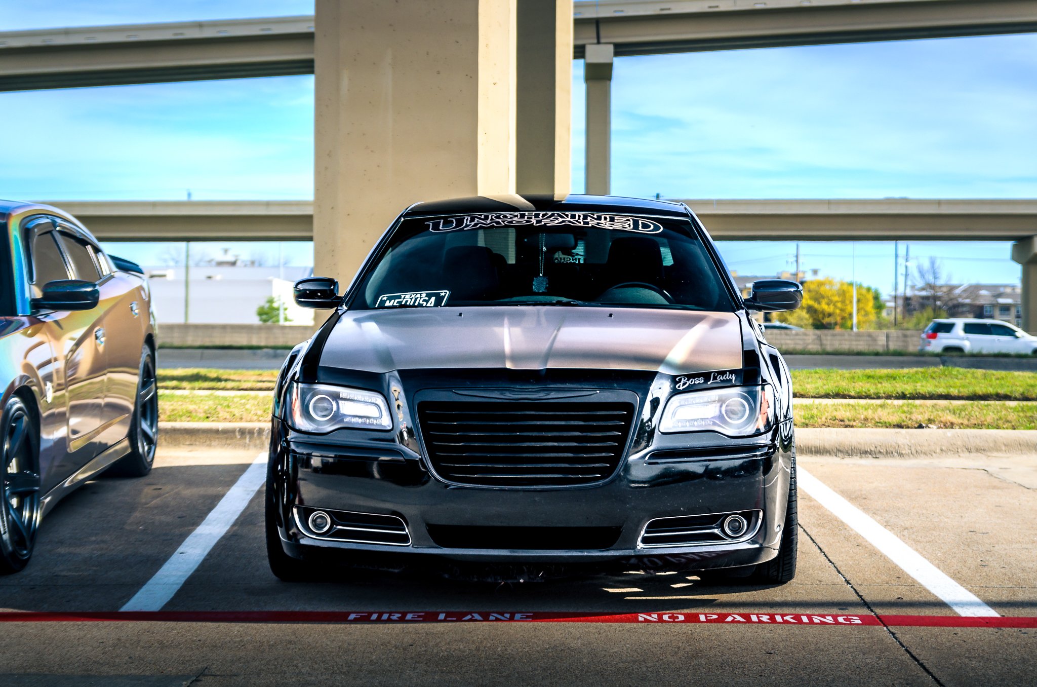 A black sedan car parked in a designated parking space in a parking lot under an overpass, with a sign on the windshield and a side decal reading 'Boss Lady'.