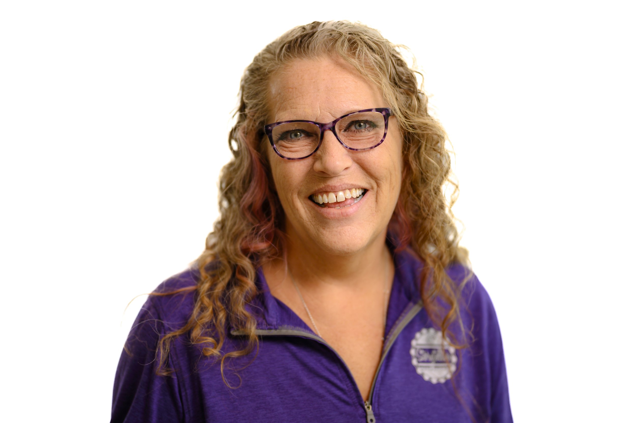 A woman with curly blonde hair wearing glasses and a purple jacket, smiling.
