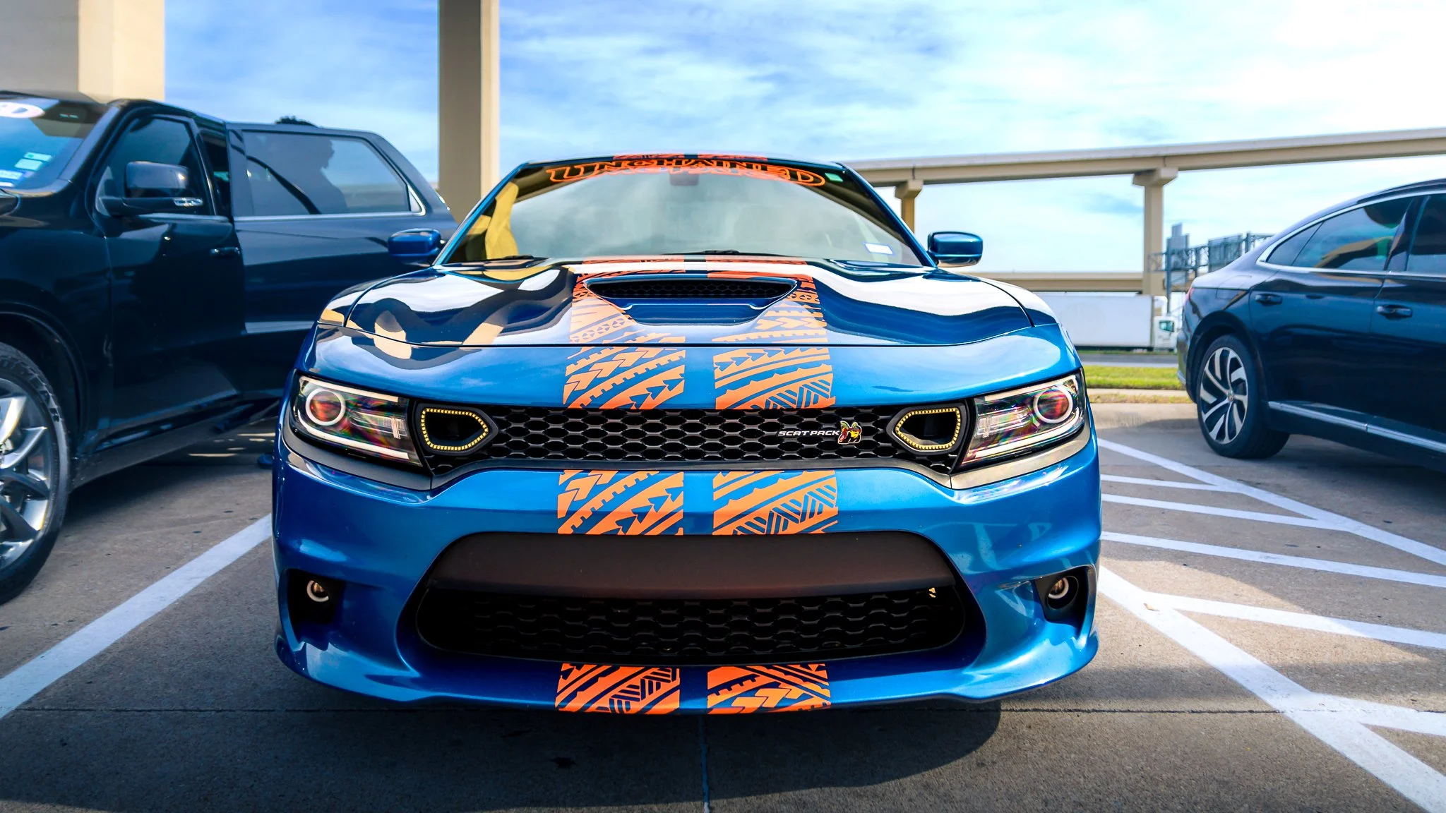 Blue sports car with an orange racing stripe parked in a parking lot, flanked by two other vehicles, beneath an elevated roadway.