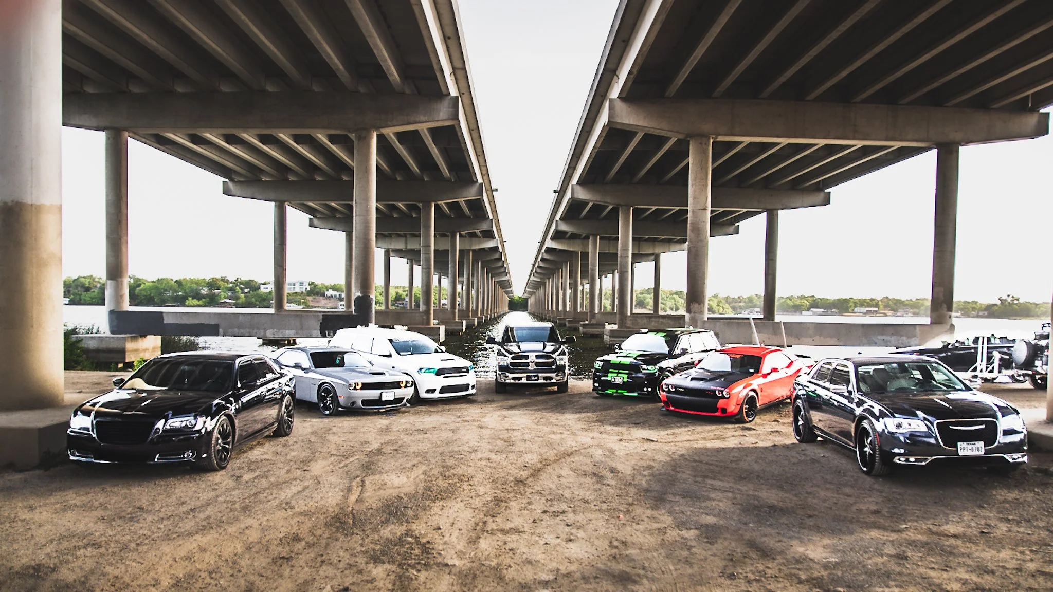 Six cars parked under a bridge near a body of water with a cityscape in the background.