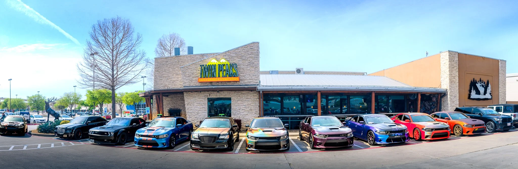 Row of customized cars parked in front of a Twin Peaks restaurant with a mountain logo, a deer statue, and leafless and leafy trees in the background on a sunny day.