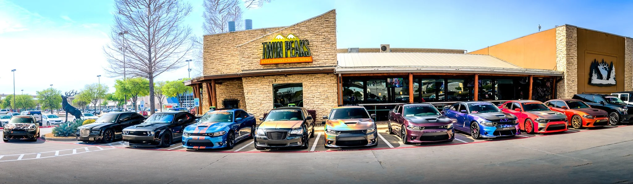 Several colorful cars parked in front of a restaurant called Twin Peaks, which has a stone facade and large windows, located in a shopping plaza with trees and parking lots visible in the background.
