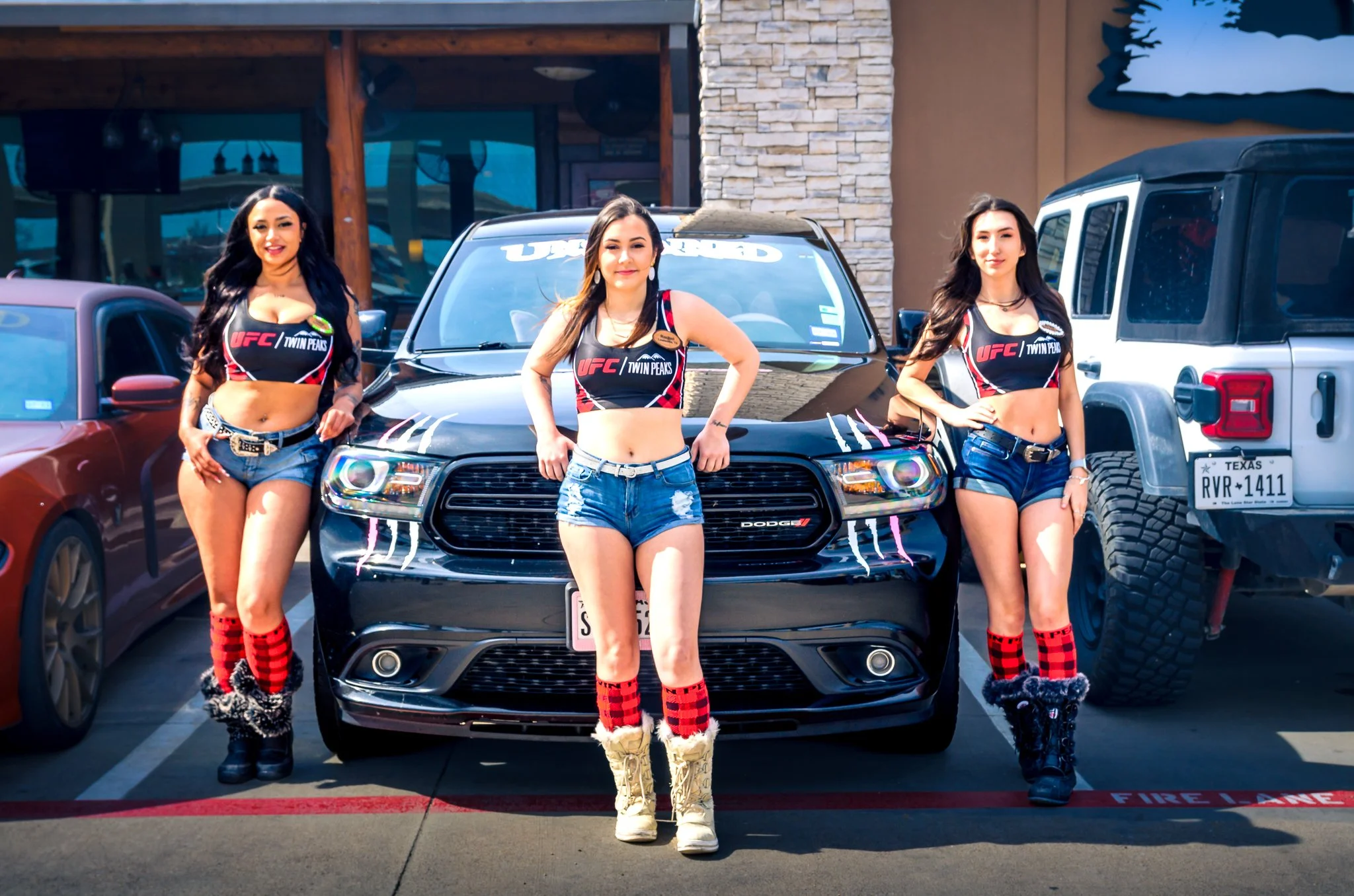 Three women dressed in UFC Twin Peaks themed clothing standing in front of a black Dodge vehicle at a parking lot, with other cars visible nearby.