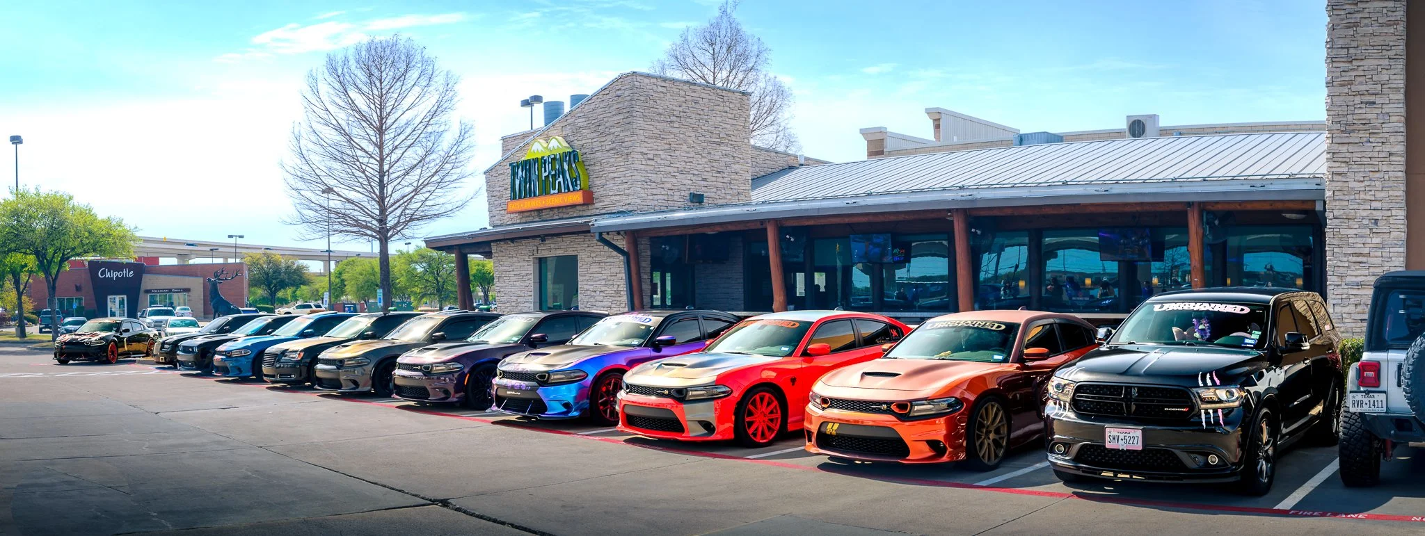 Line of colorful sporty cars parked outside a restaurant called Twisted Tykes, with a Chipotle nearby and a large deer sculpture visible.