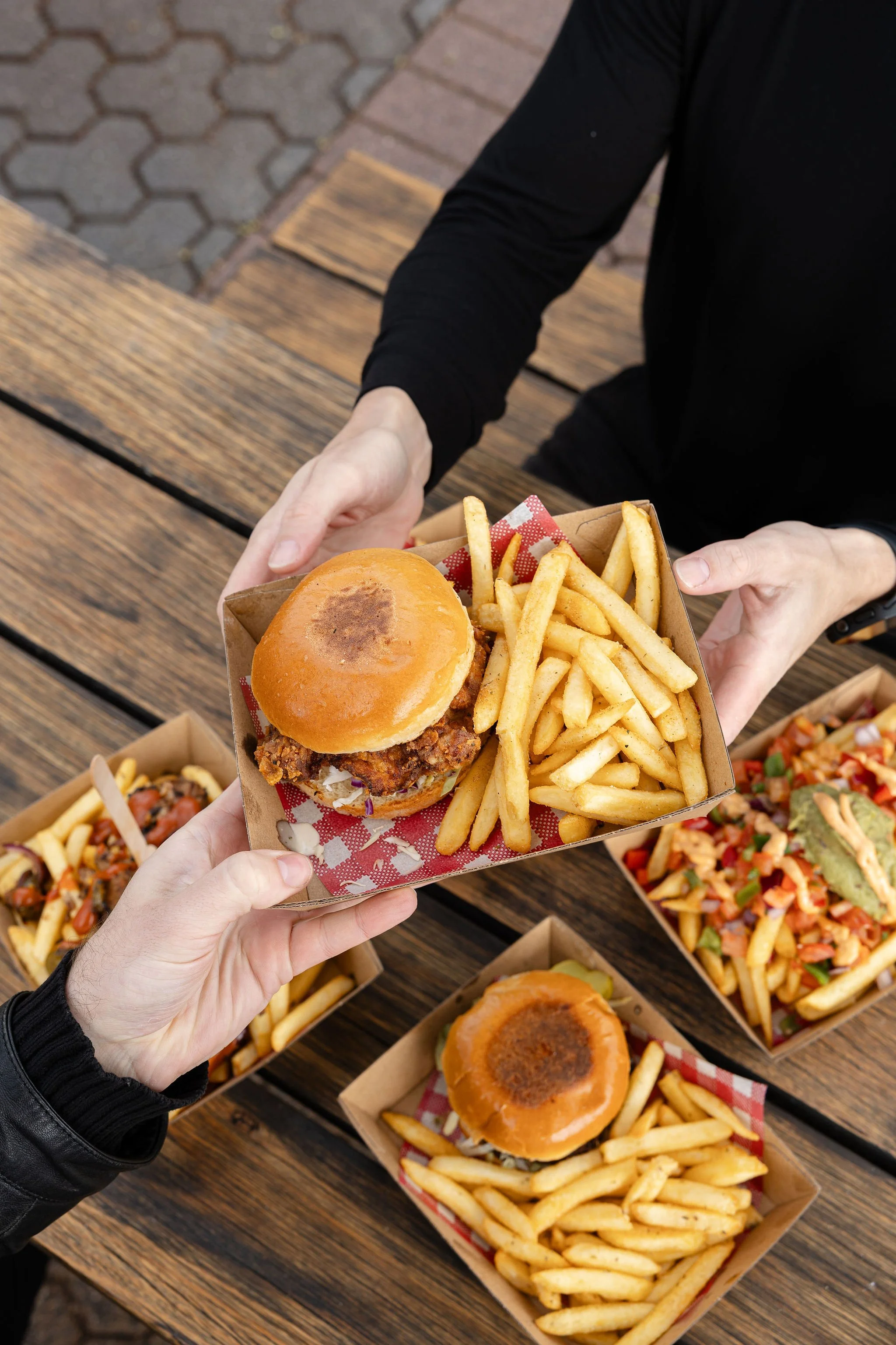 A person in a black jacket holding a tray with a fried chicken sandwich and French fries, at an outdoor wooden table with several other food containers and a brick pavement in the background.