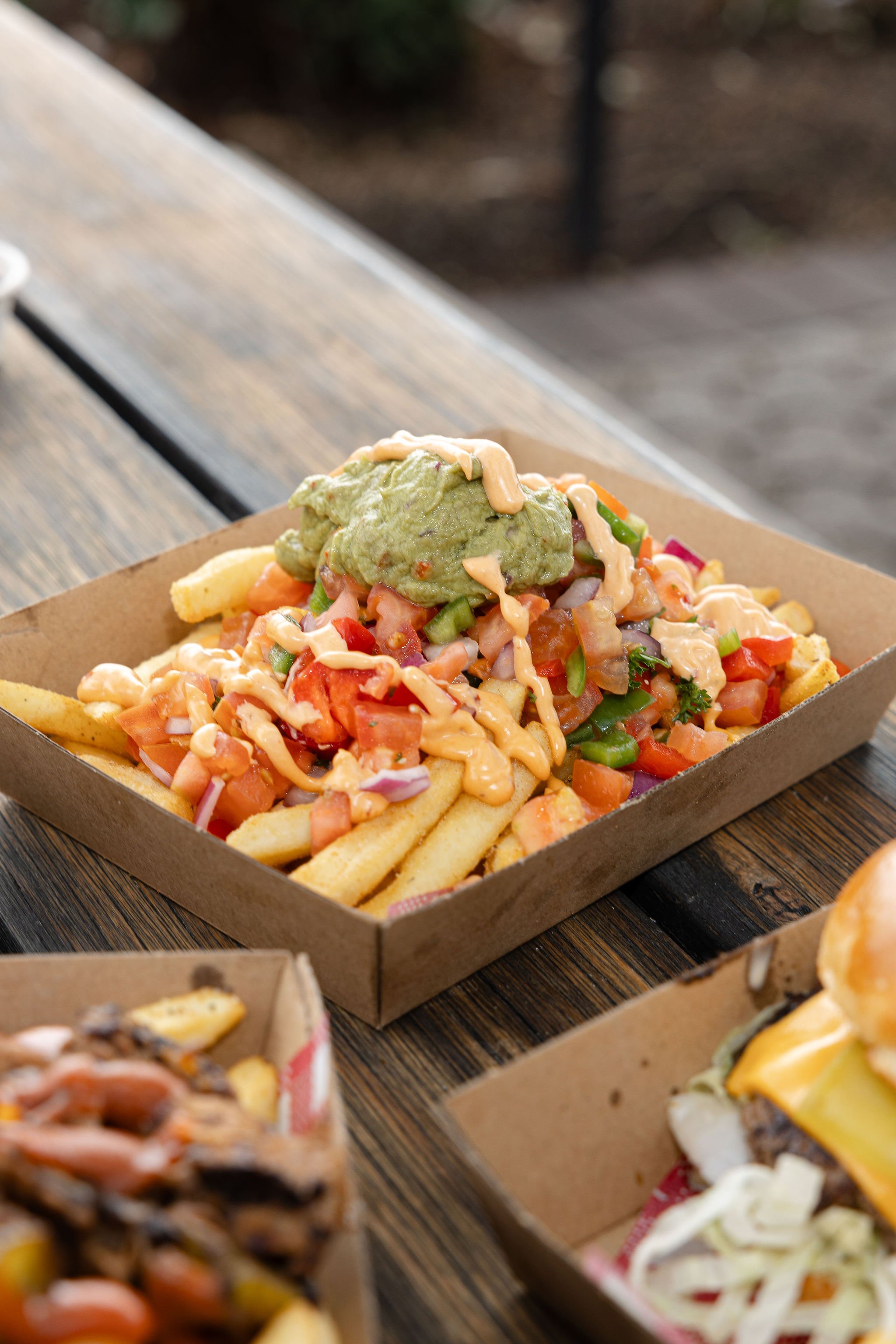 Close-up of a tray of loaded fries topped with guacamole, diced tomatoes, green peppers, chopped onions, creamy dressing, and herbs, set on a wooden table.