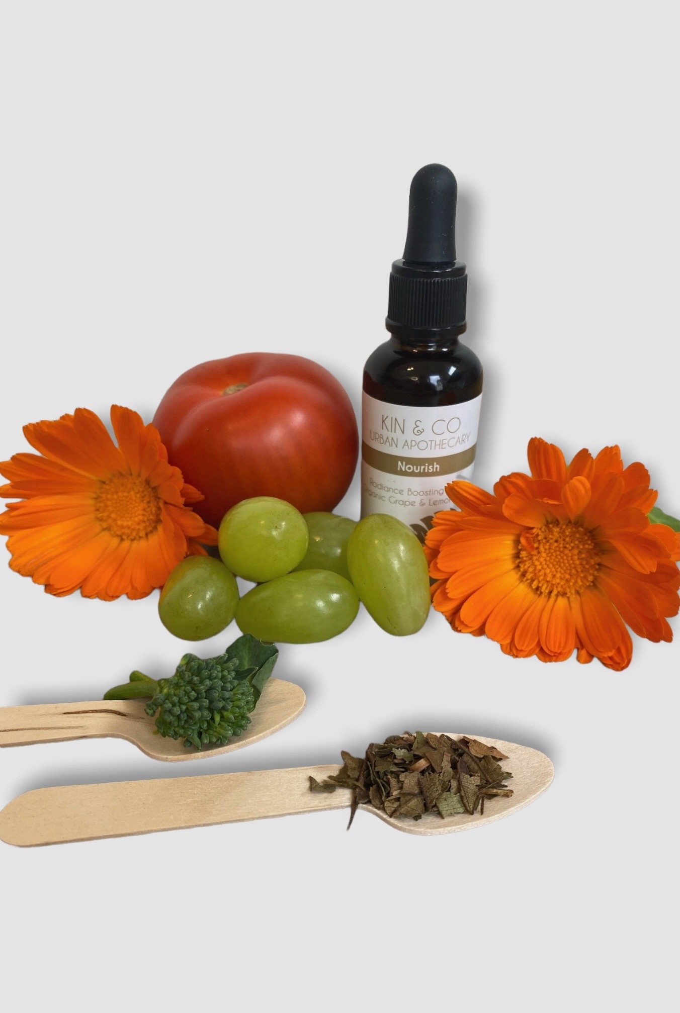 Nourish Radiance Boosting Oil Blend 30 ml Bottle surround by grapes, a tomato and calendula flowers, with two wooden spoons, one containing broccoli the other Lemon Myrtle.
