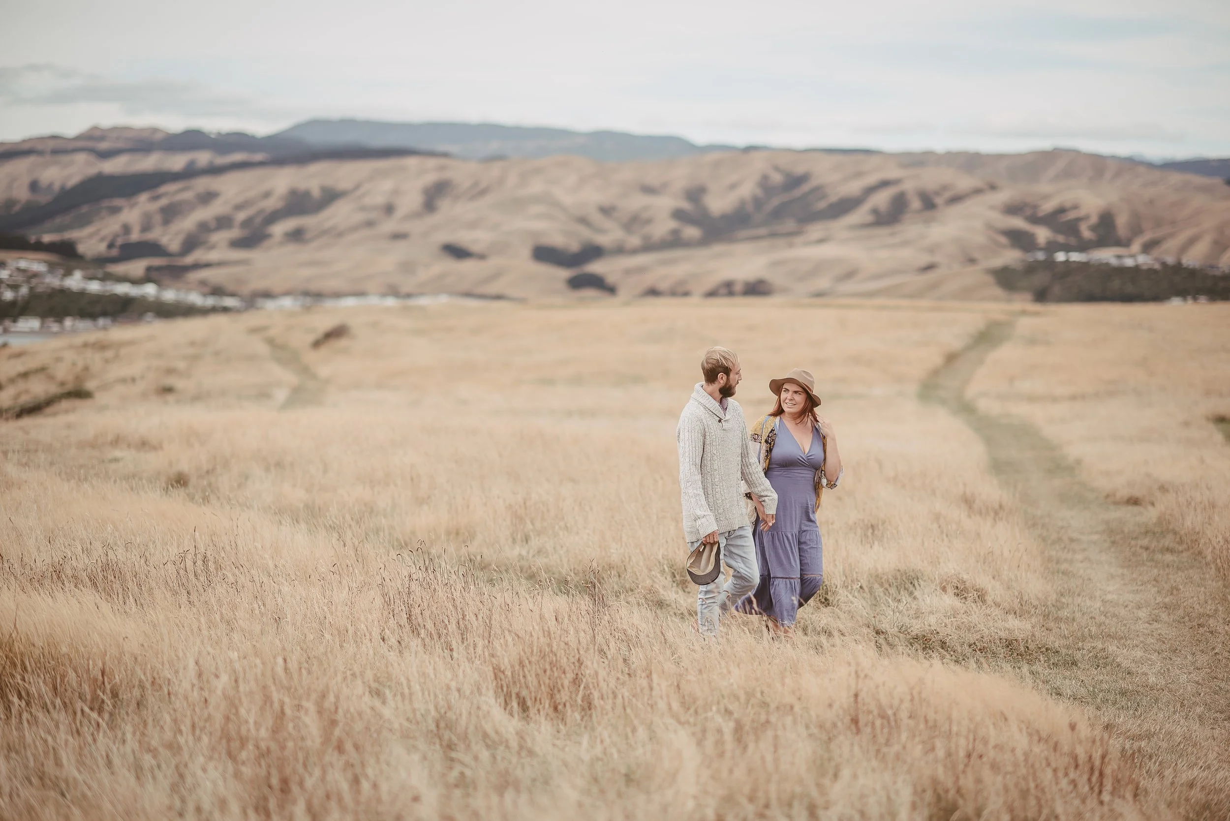 A man and a woman holding hands and walking through a grassy field with hills in the background.
