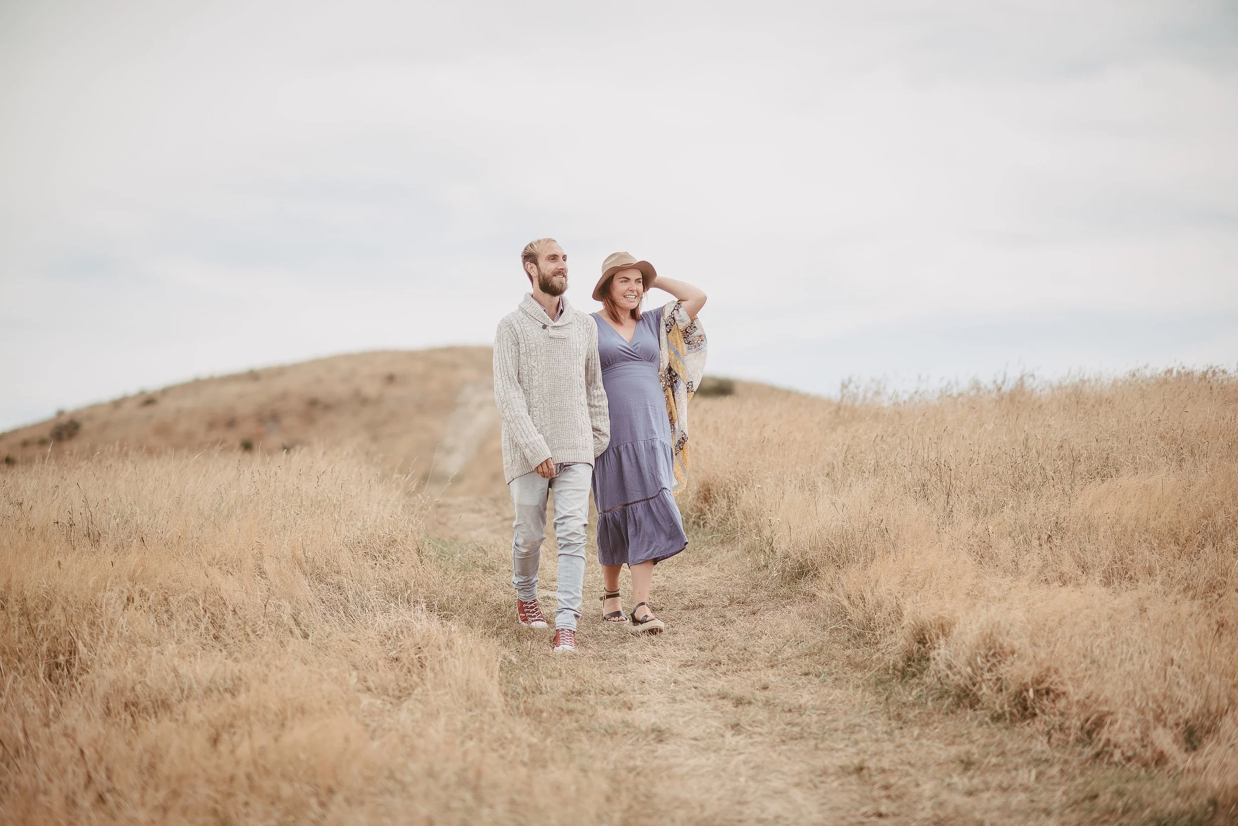 A man and woman walking together in a field of tall, golden grass under a cloudy sky, smiling and enjoying the moment. The woman is wearing a hat and a long dress, the man is in a sweater and jeans.