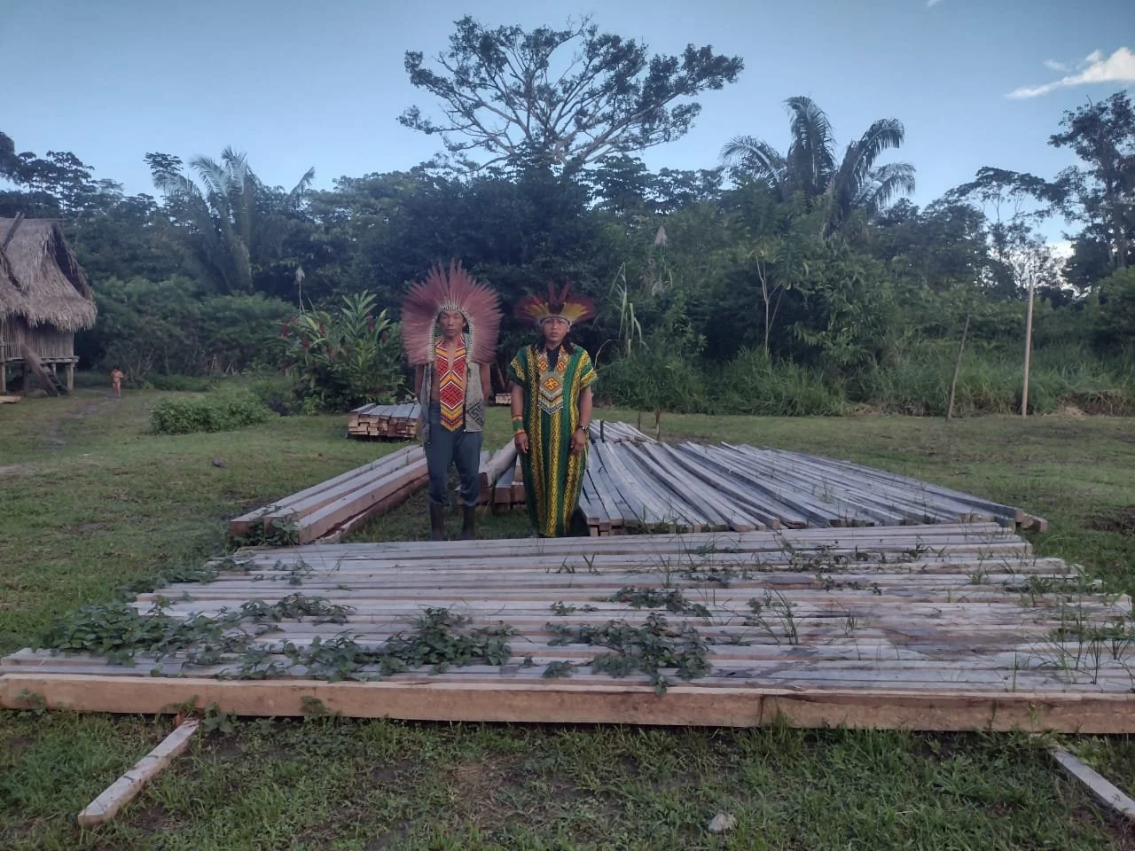 Two indigenous people wearing traditional headdresses and clothing standing on a grassy field with a construction site of a wooden structure in front. There are trees and huts in the background.